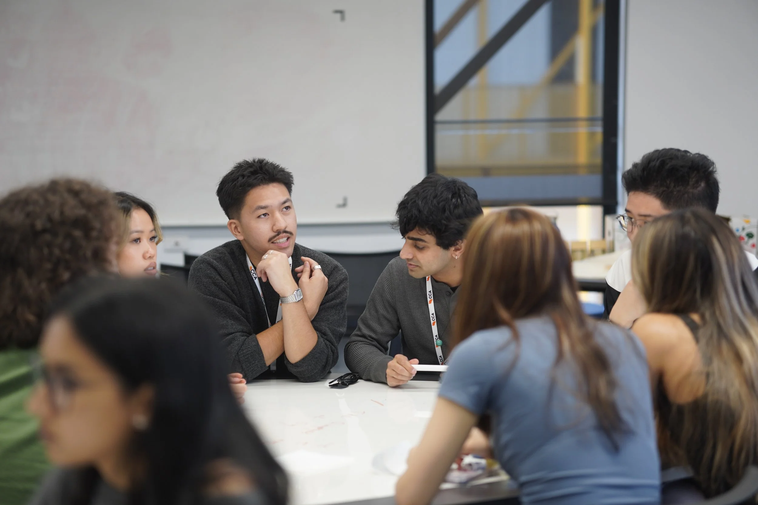 A group of diverse young adults sitting around a table in a classroom or conference room, engaged in a discussion or conversation.