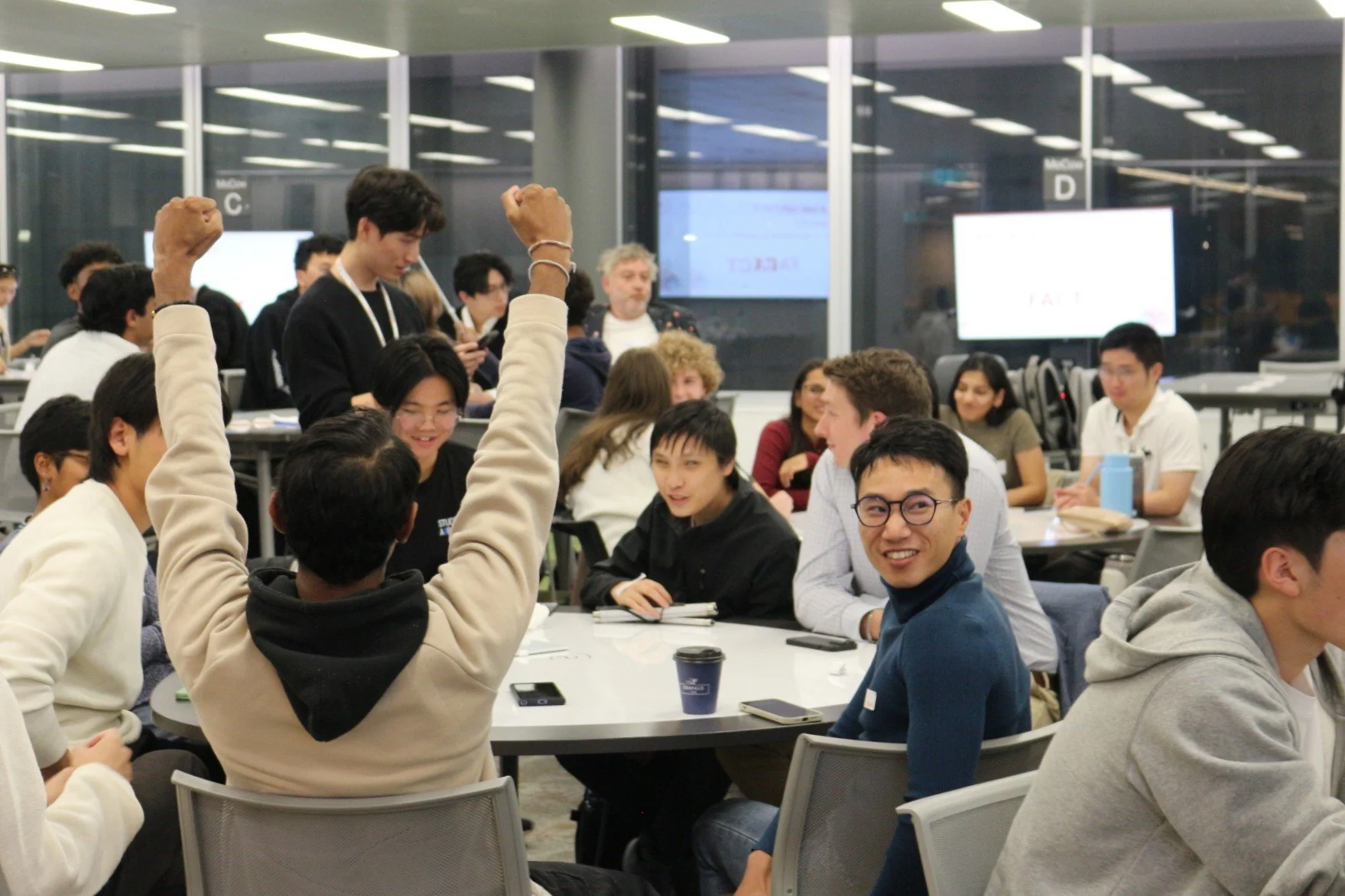 A diverse group of people seated around tables in a modern conference room, with some raising hands and others engaging in conversation or listening attentively.