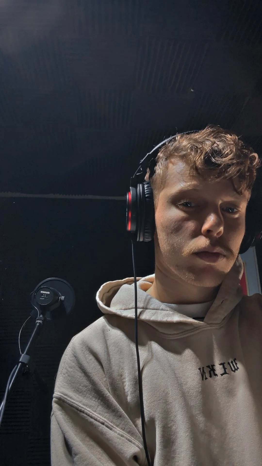 A young man with curly hair and a beige hoodie, wearing headphones, taking a selfie in a recording studio with dark soundproof walls and a microphone in the background.