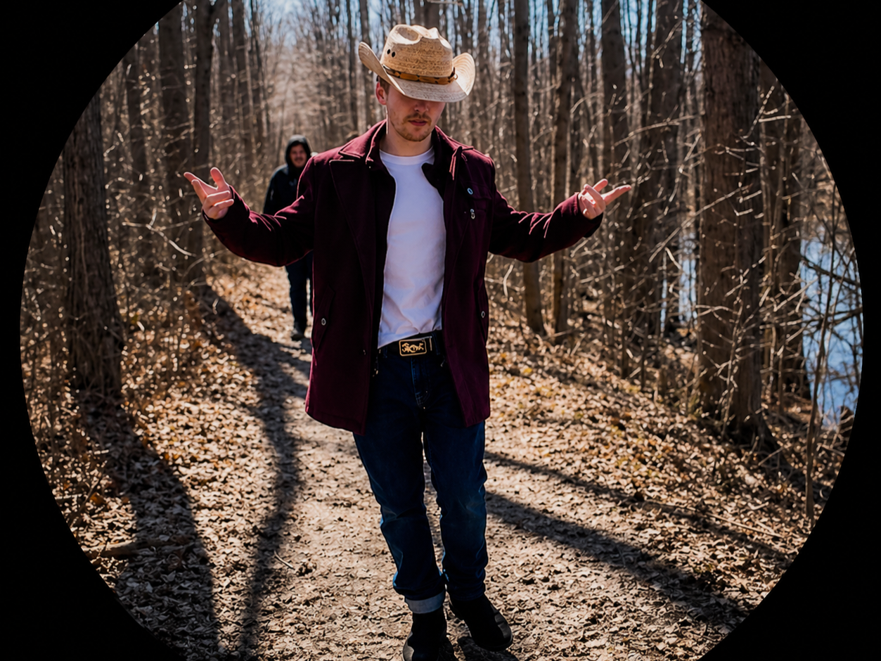 A man wearing a cowboy hat, burgundy jacket, white shirt, and jeans walks on a forest trail with another man in the background. The forest has leafless trees and a small body of water visible to the right.