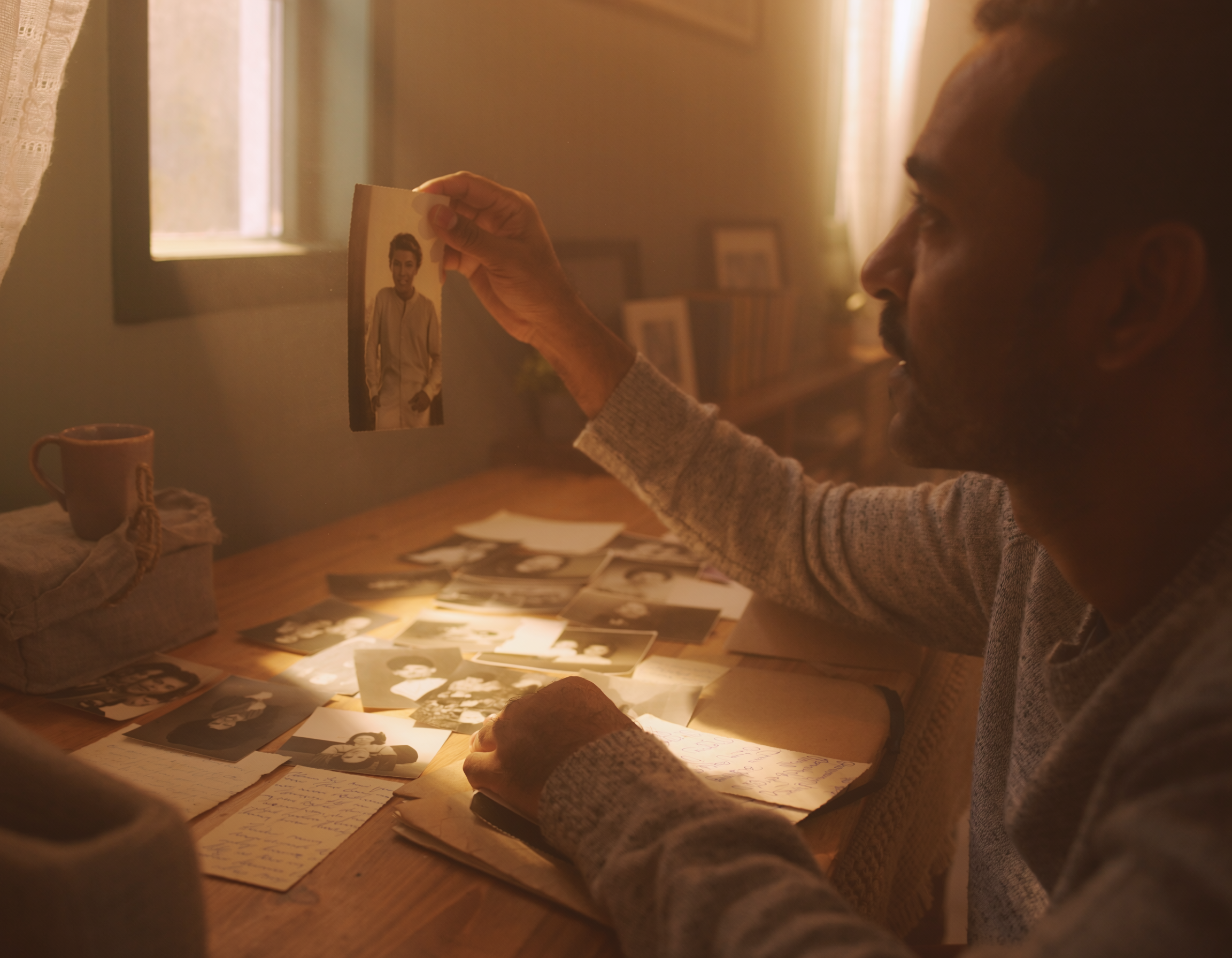 A person holding a black-and-white photograph of a woman while sitting at a wooden table covered with various photographs and handwritten letters. There is warm sunlight coming through a window.