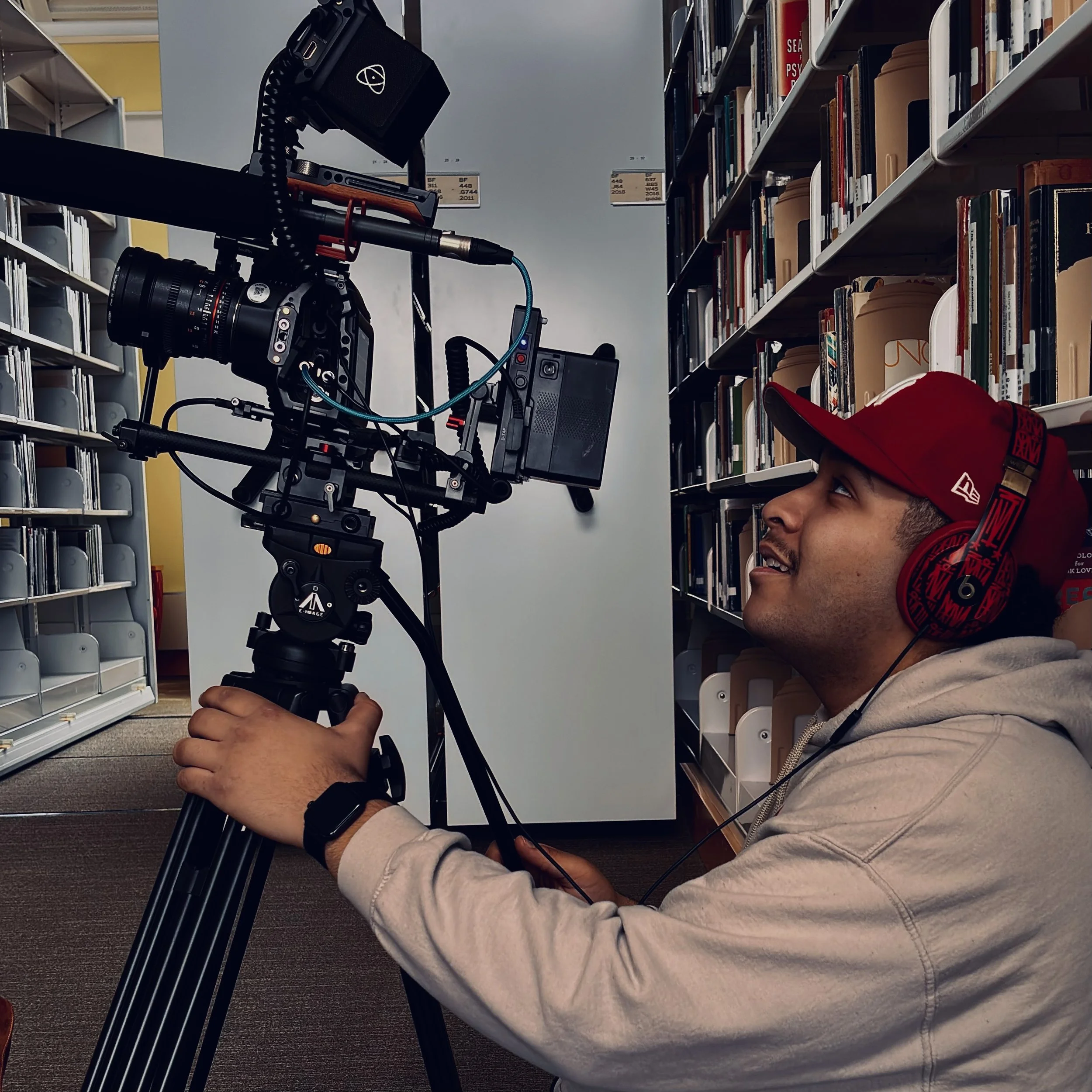A person wearing headphones operating a professional video camera on a tripod in a library aisle.