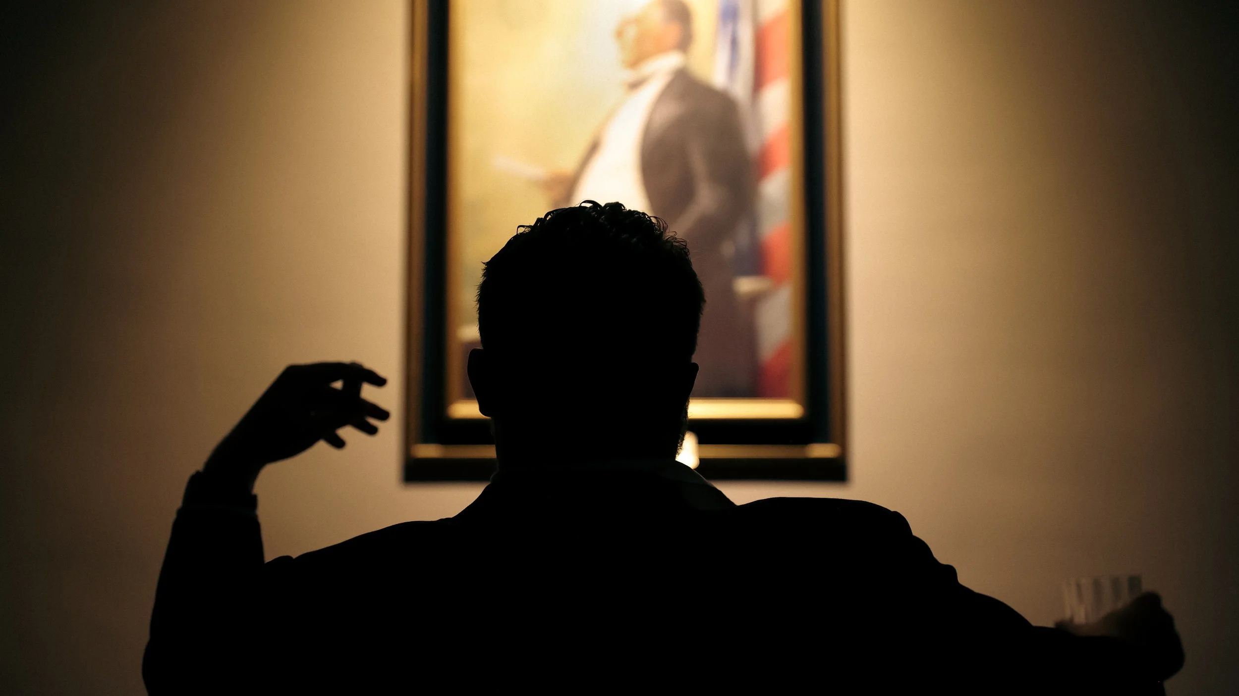 A silhouette of a person with curly hair sitting in front of a framed portrait of a man in a suit and glasses.