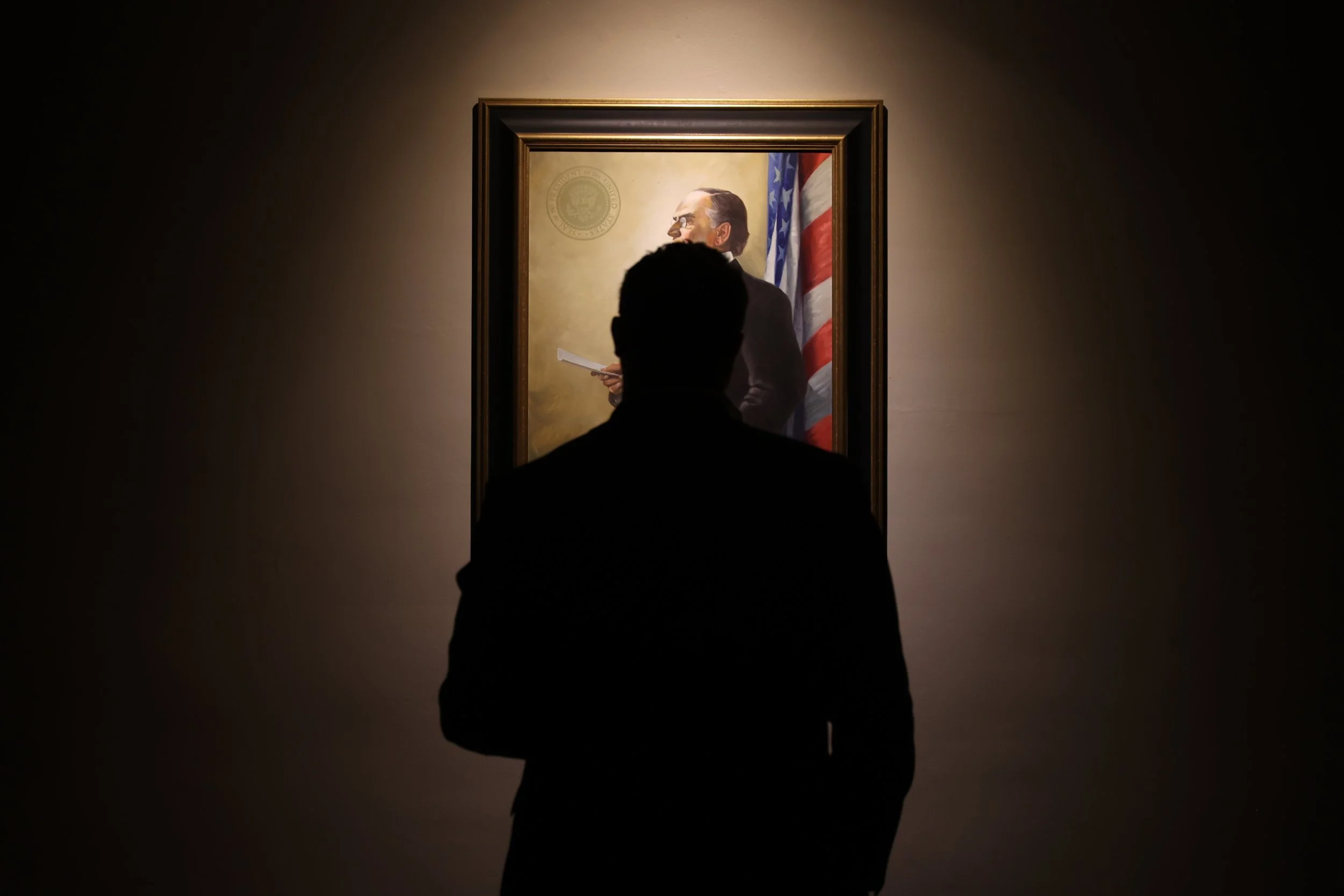 Person standing in front of a framed portrait on a wall, with an American flag behind the portrait.