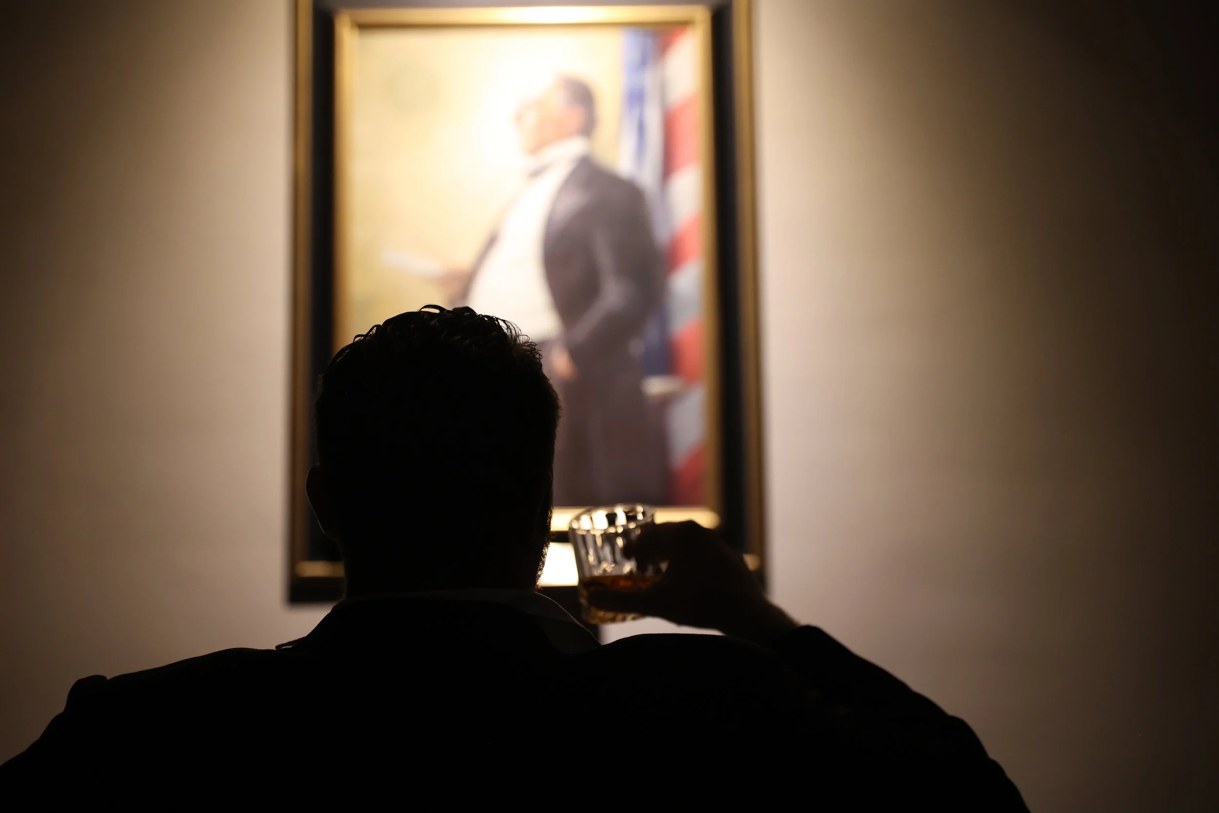 A person sitting in a room, facing a mirror with a blurred image of a man in a tuxedo and American flag in the background.