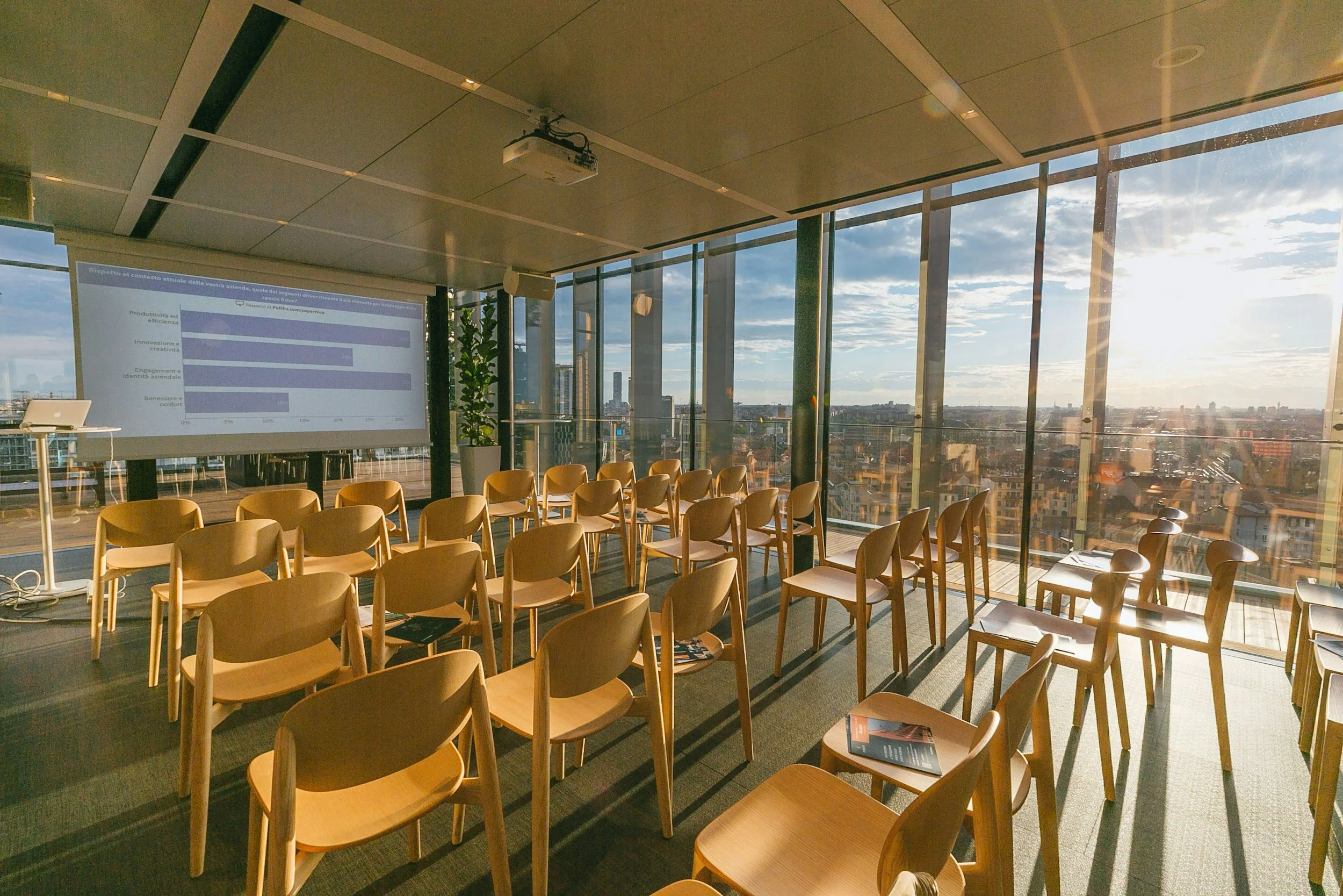 A conference room with large windows showing a cityscape and a partly cloudy sky. The room has rows of wooden chairs facing a screen, and a projector is mounted on the ceiling. The sunlight creates a warm glow inside the room.