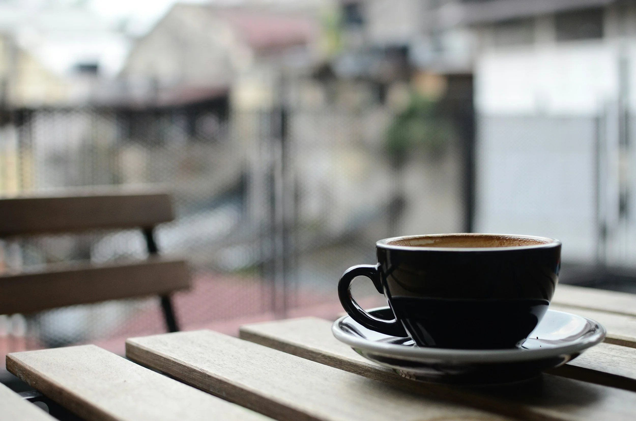 Black coffee cup on a saucer placed on a wooden table indoors, with blurred background of a balcony or patio.