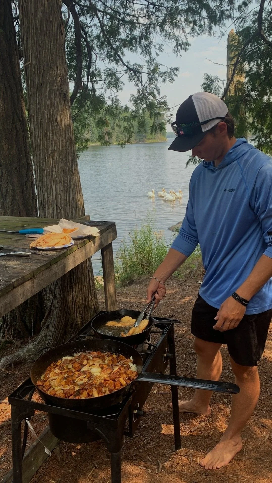 A person cooking food on a portable stove outdoors near a lake, with swans swimming in the water in the background, under trees.