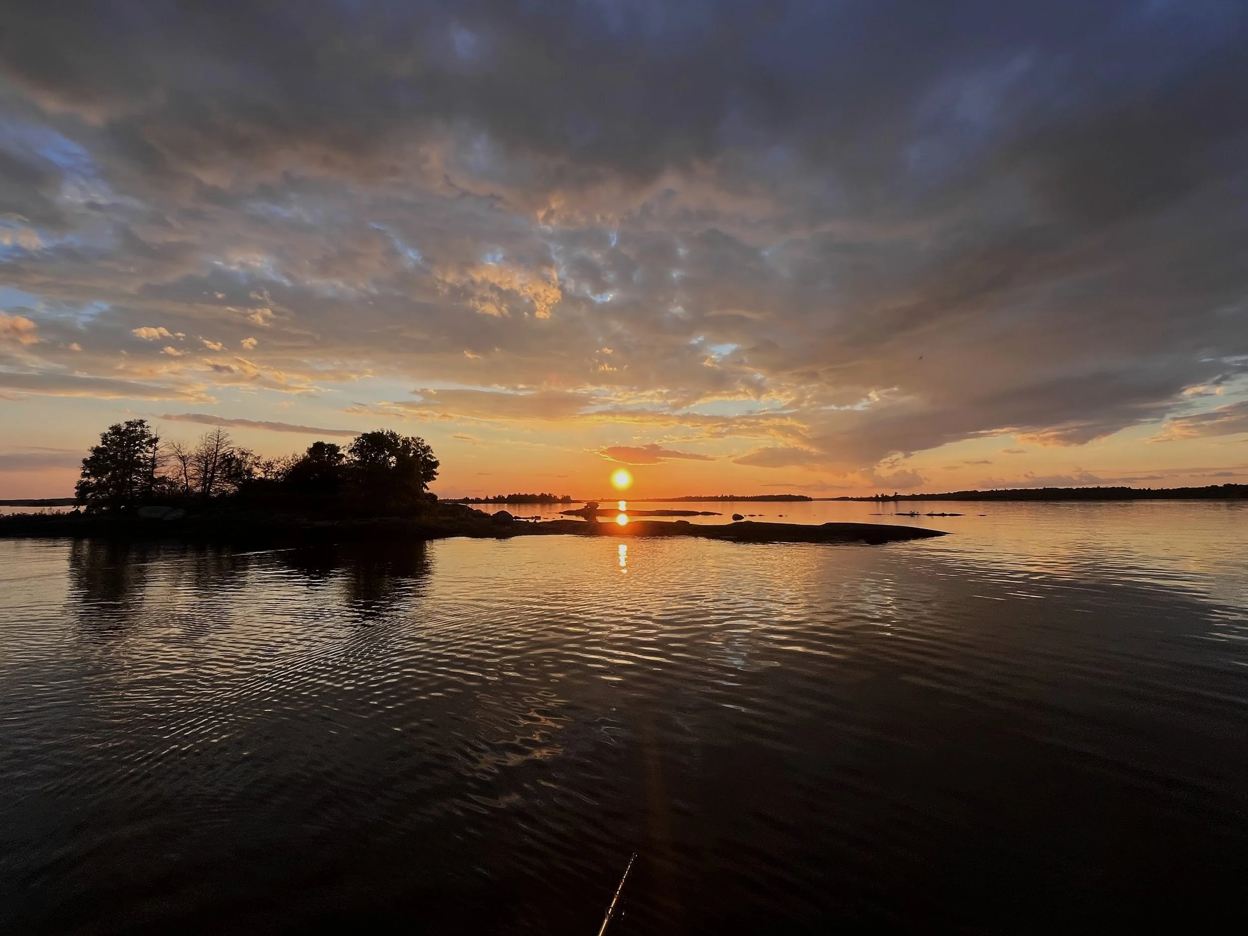 Sunset over a body of water with trees in the foreground and a partly cloudy sky.