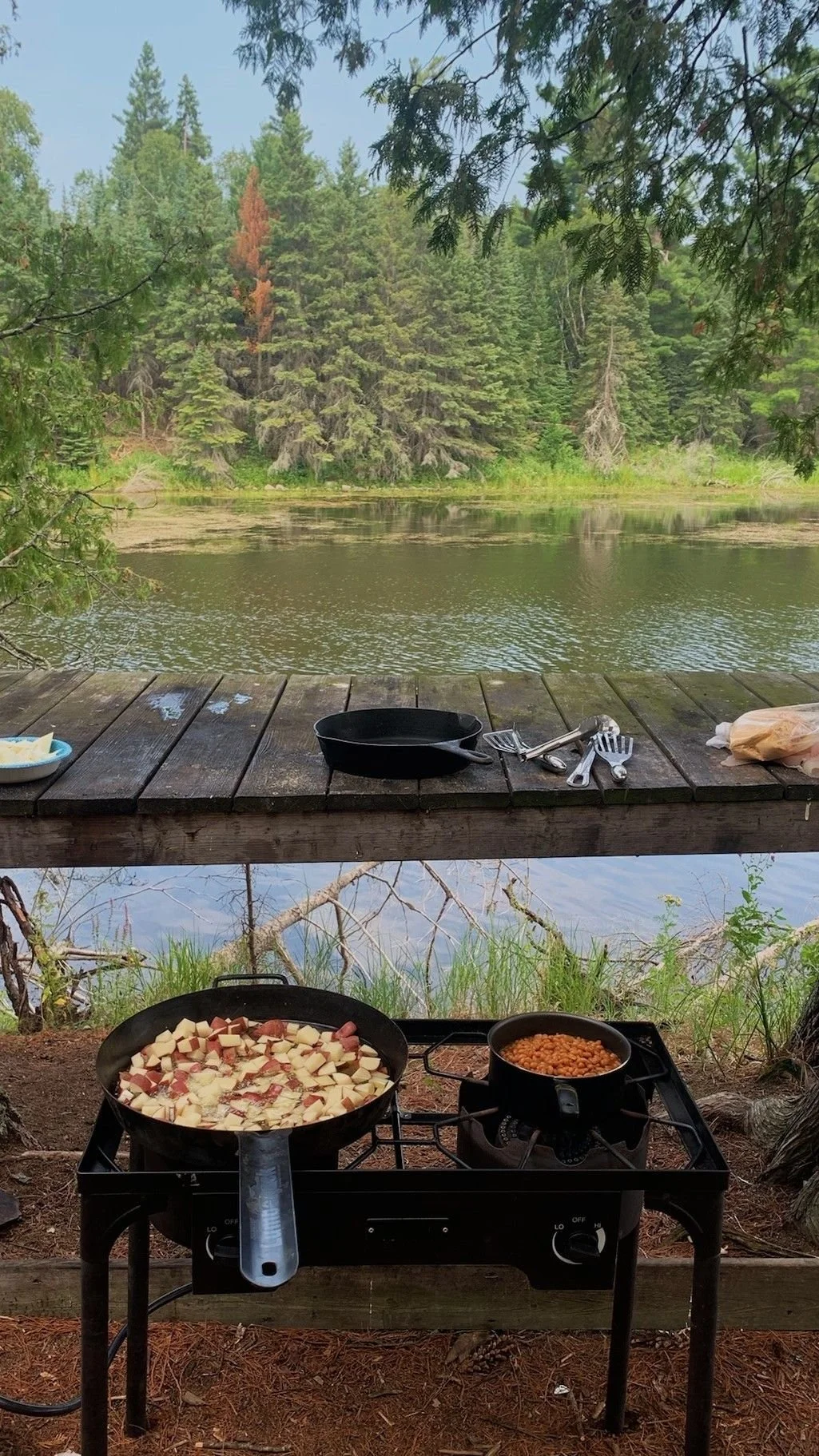 Cooking food over a portable stove by a lakeside, with a wooden deck, a frying pan with diced potatoes and bacon, and a pot of baked beans, surrounded by trees and water.