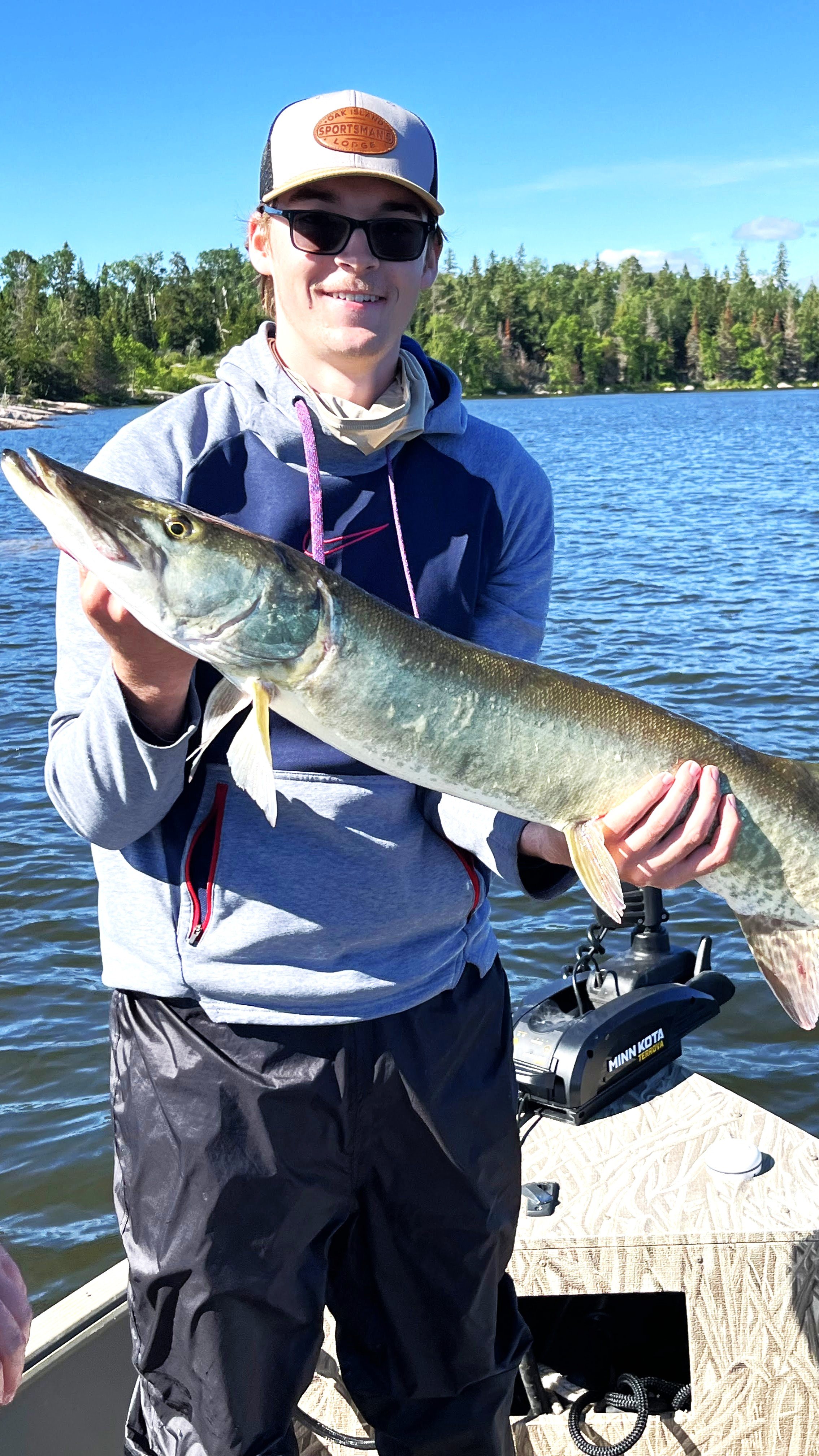 A young man wearing a gray hoodie, sunglasses, and a hat holding a large fish on a boat on a lake with trees in the background.