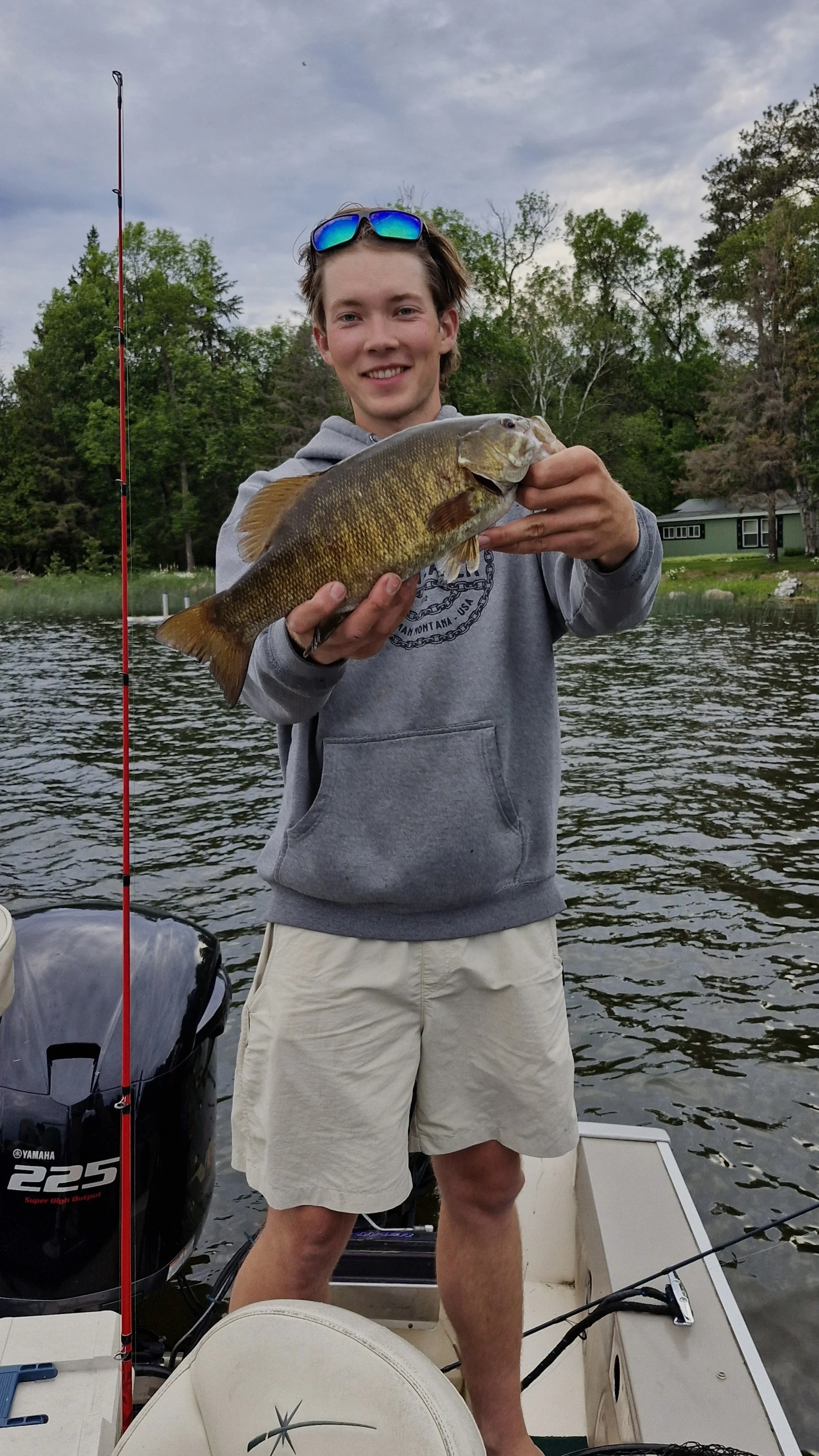 A young man smiling and holding a large fish on a boat on a lake with trees and cloudy sky in the background.