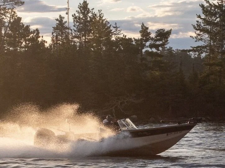 A motorboat speeding across a lake or river, leaving a spray of water behind, with trees and a cloudy sky in the background.