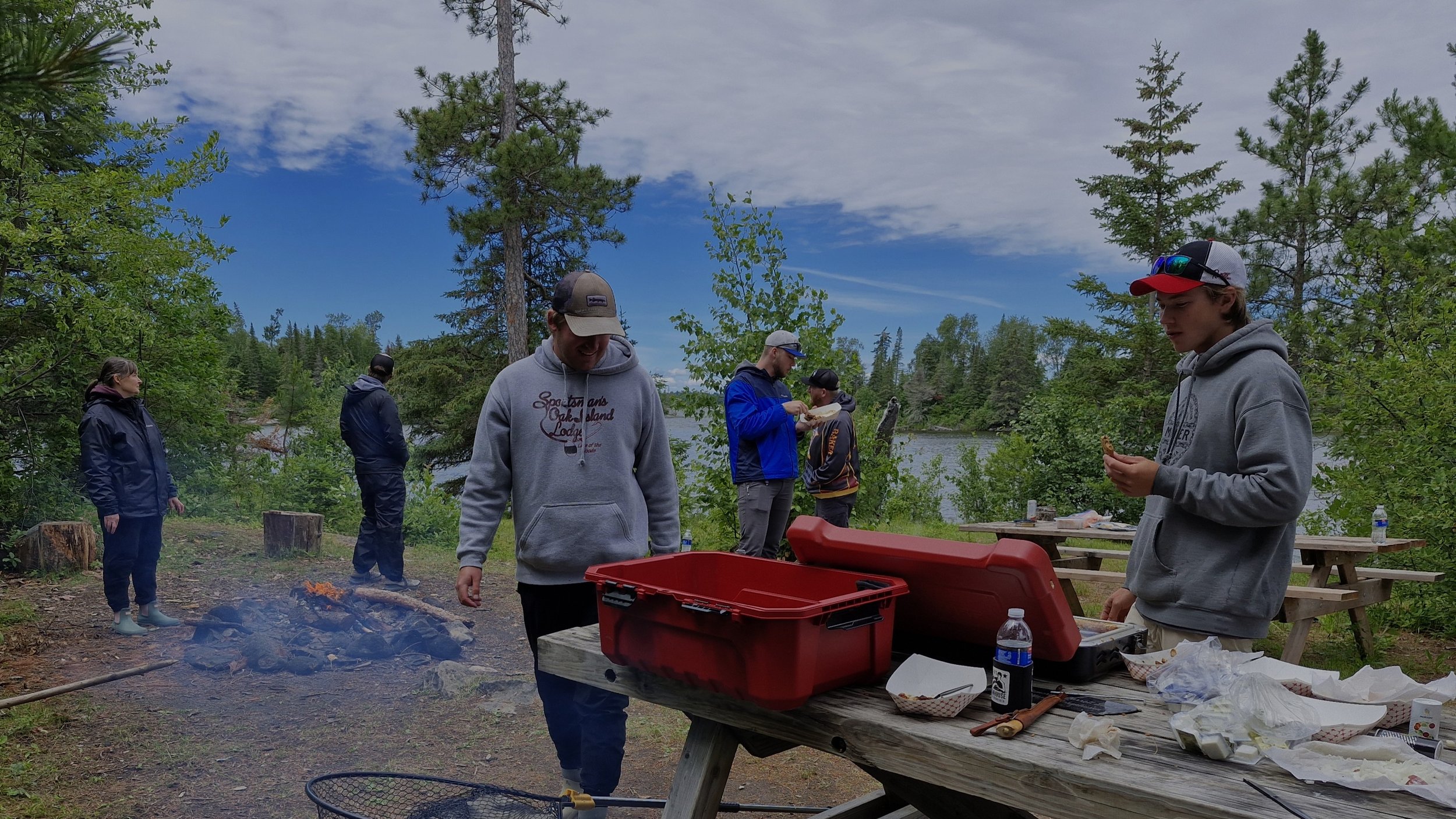 Group of people outdoors near a river, standing around a picnic table and a campfire, surrounded by trees and cloudy sky.