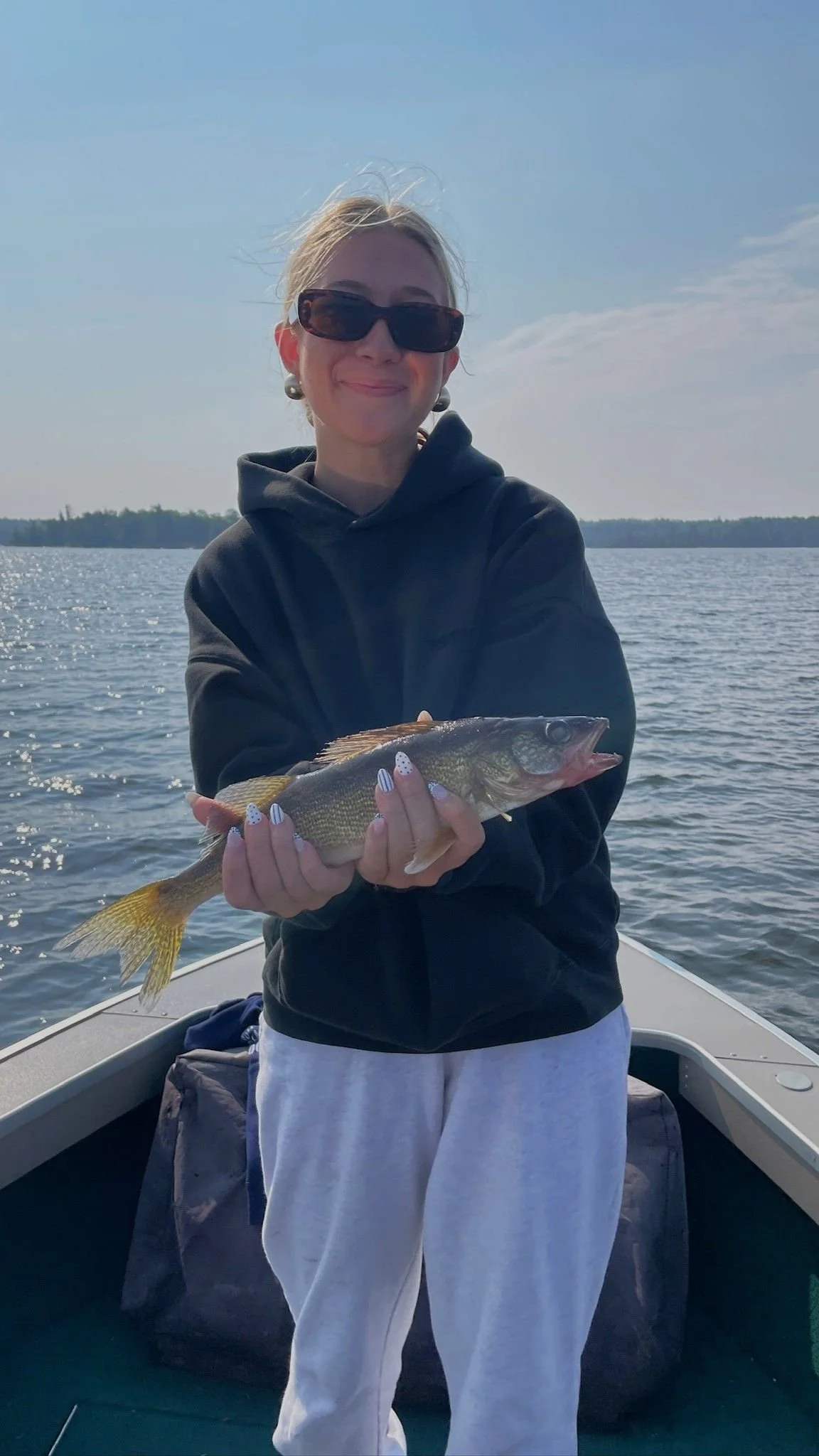 A woman smiling and wearing sunglasses, holding a fish on a boat in a lake with a clear sky in the background.