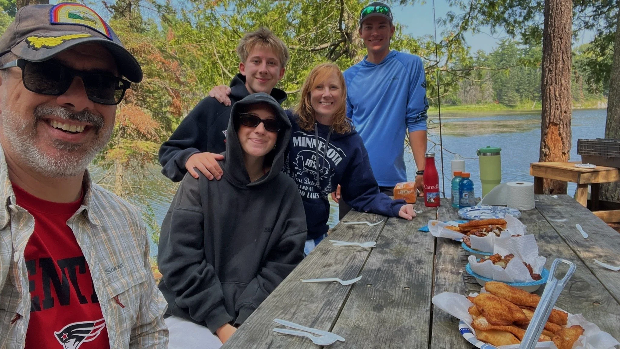 Group of six people, smiling, gathered around a picnic table outdoors near a lake, with food and drinks on the table, trees and water in the background.