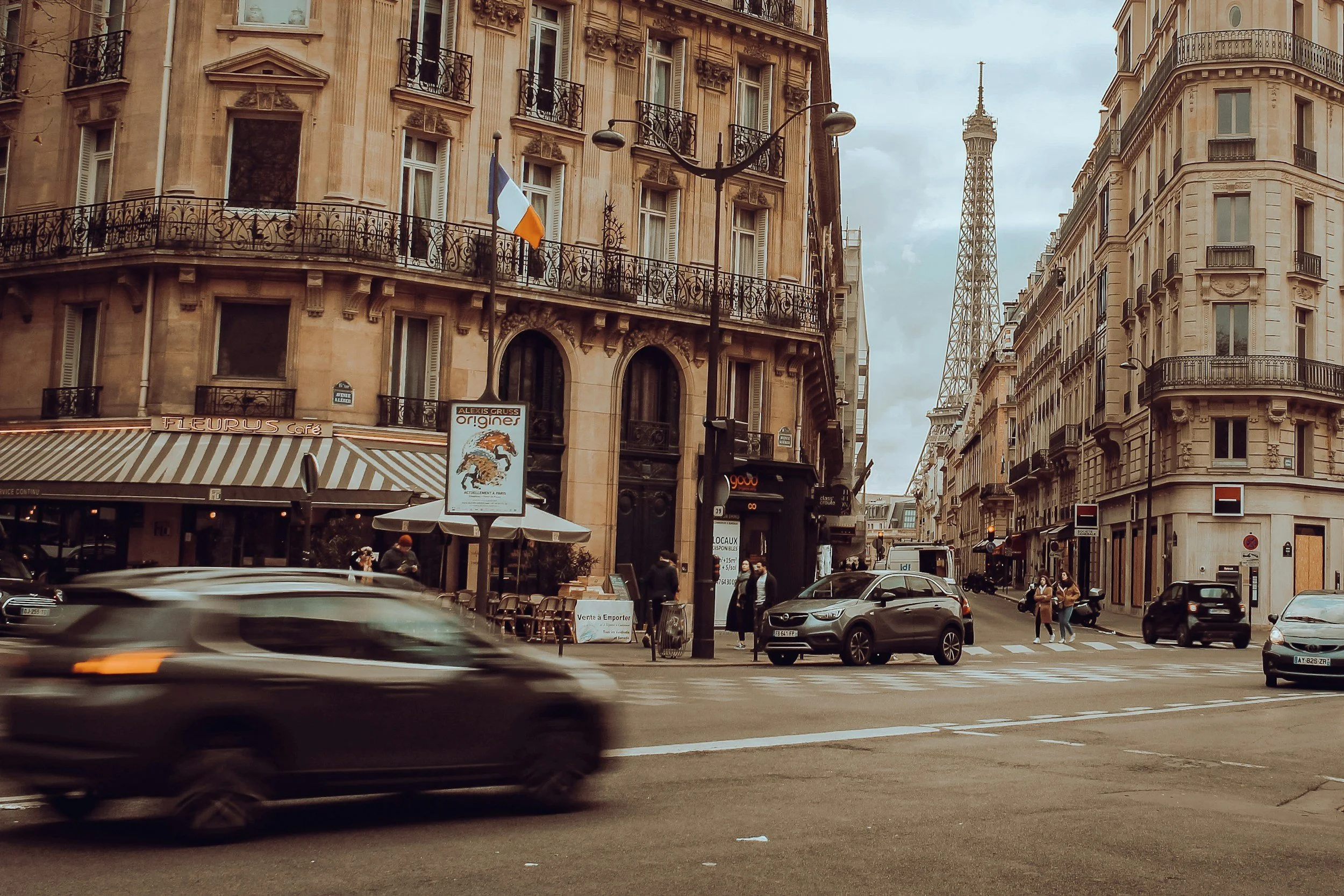 Parisian street scene with classic Haussmann-style buildings, a partial view of the Eiffel Tower in the background, traffic on the road, pedestrians crossing, and storefront signs including a cafe and a sign for Alexis Gruss origins.