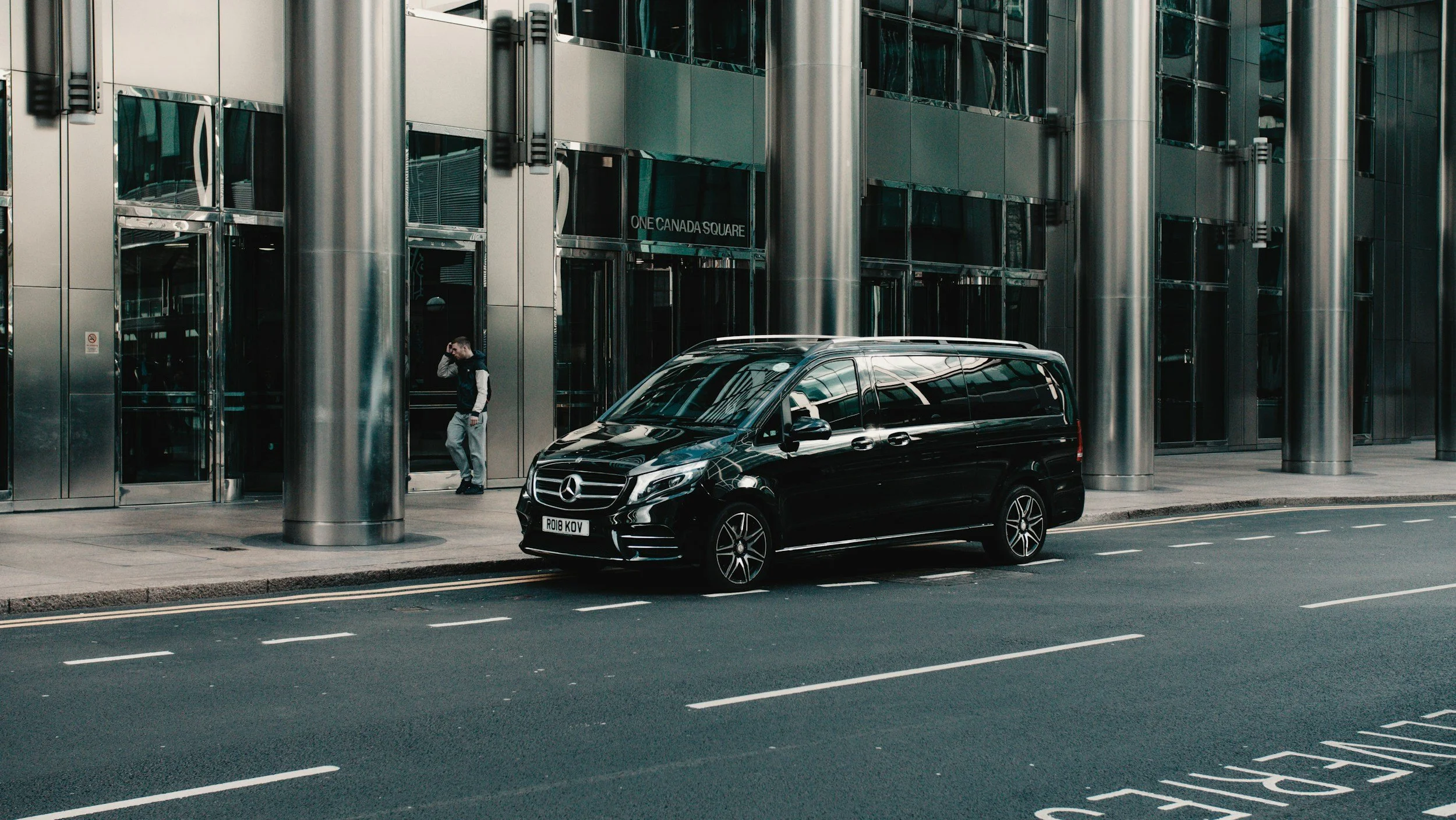 A black Mercedes-Benz van parked on the street outside a modern glass and metal office building. A man is walking past the entrance, talking on a mobile phone. The building has a sign that reads 'ONE CANADA SQUARE' and large cylindrical metal columns.
