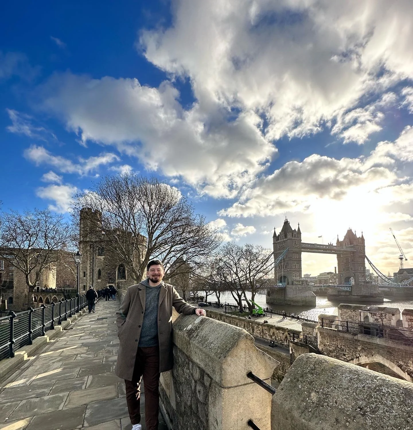 A man smiling and standing on a bridge in front of Tower Bridge in London, with trees and historic buildings on the riverbank and a partly cloudy sky overhead.