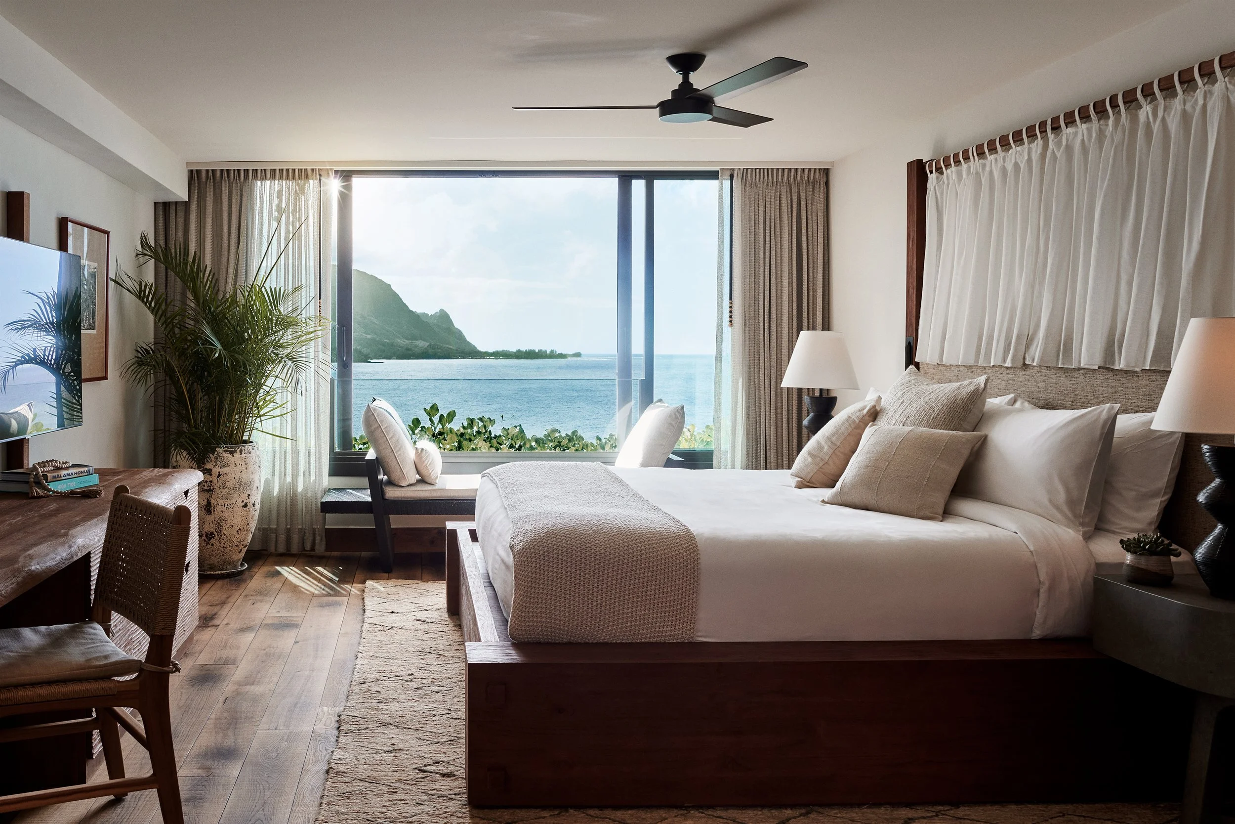A bedroom with a large window overlooking the ocean and mountains, decorated with neutral tones, a bed with pillows, a nightstand with a lamp, potted plants, and curtains.