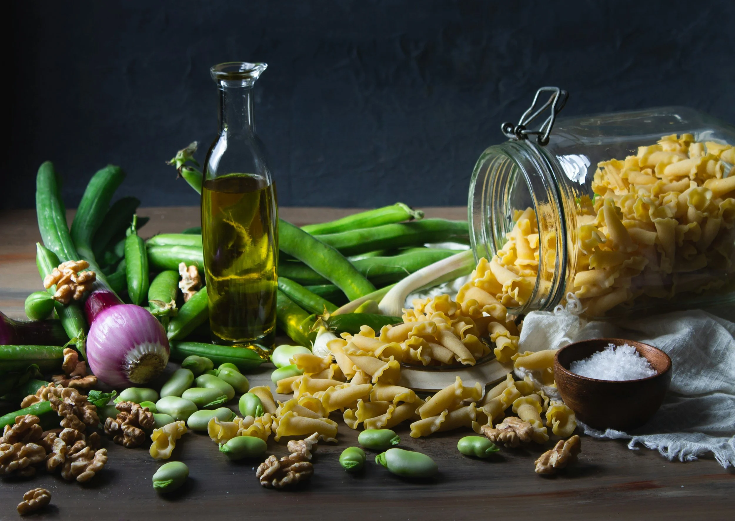 Assorted ingredients for pasta or salad on a wooden surface, including green beans, walnuts, red onion, pasta, olive oil in a bottle, coarse salt in a bowl, and a glass jar of pasta. The background is dark and minimalist.