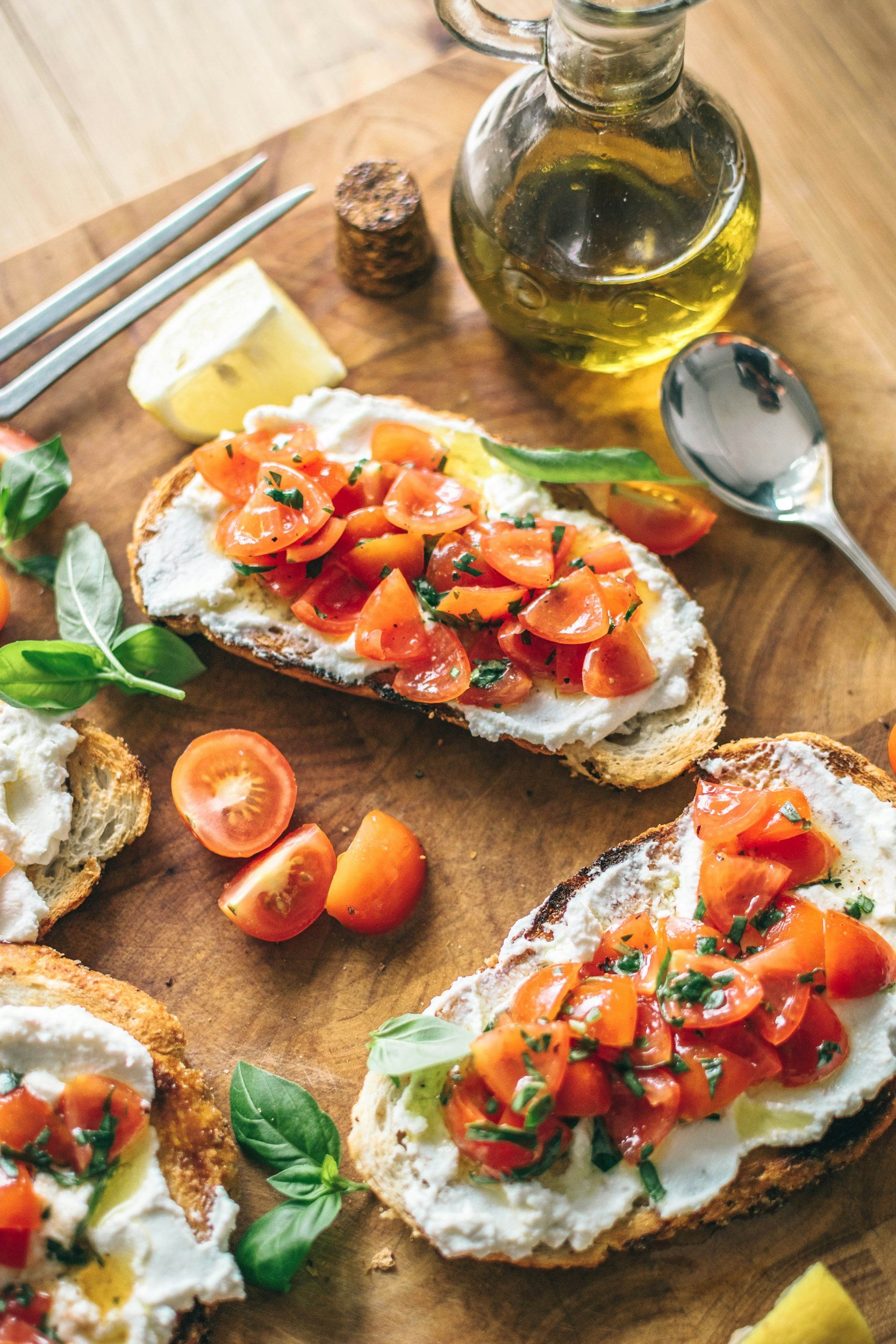 Open-faced bread with cream cheese, cherry tomato slices, and fresh basil on a wooden cutting board with a wedge of lemon, a bottle of olive oil, a cork, and a spoon.