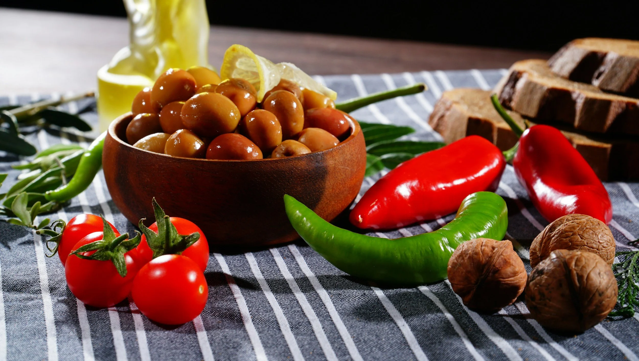 A bowl of green olives garnished with lemon slices, surrounded by tomatoes, green and red chili peppers, walnuts, and pieces of bread on a striped cloth.