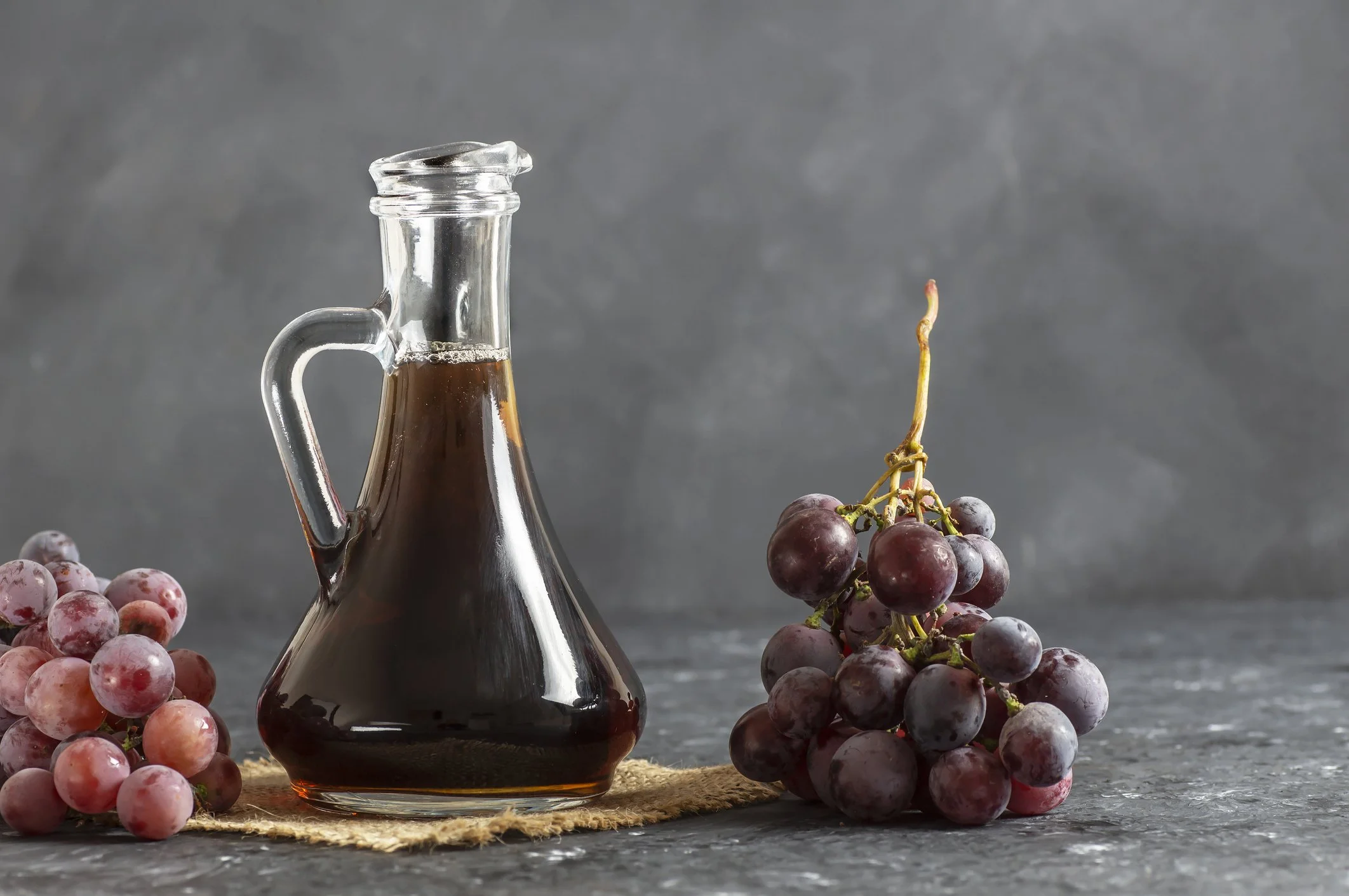 A glass bottle of red wine with a glass stopper, surrounded by bunches of red grapes on a cork placemat against a gray background.