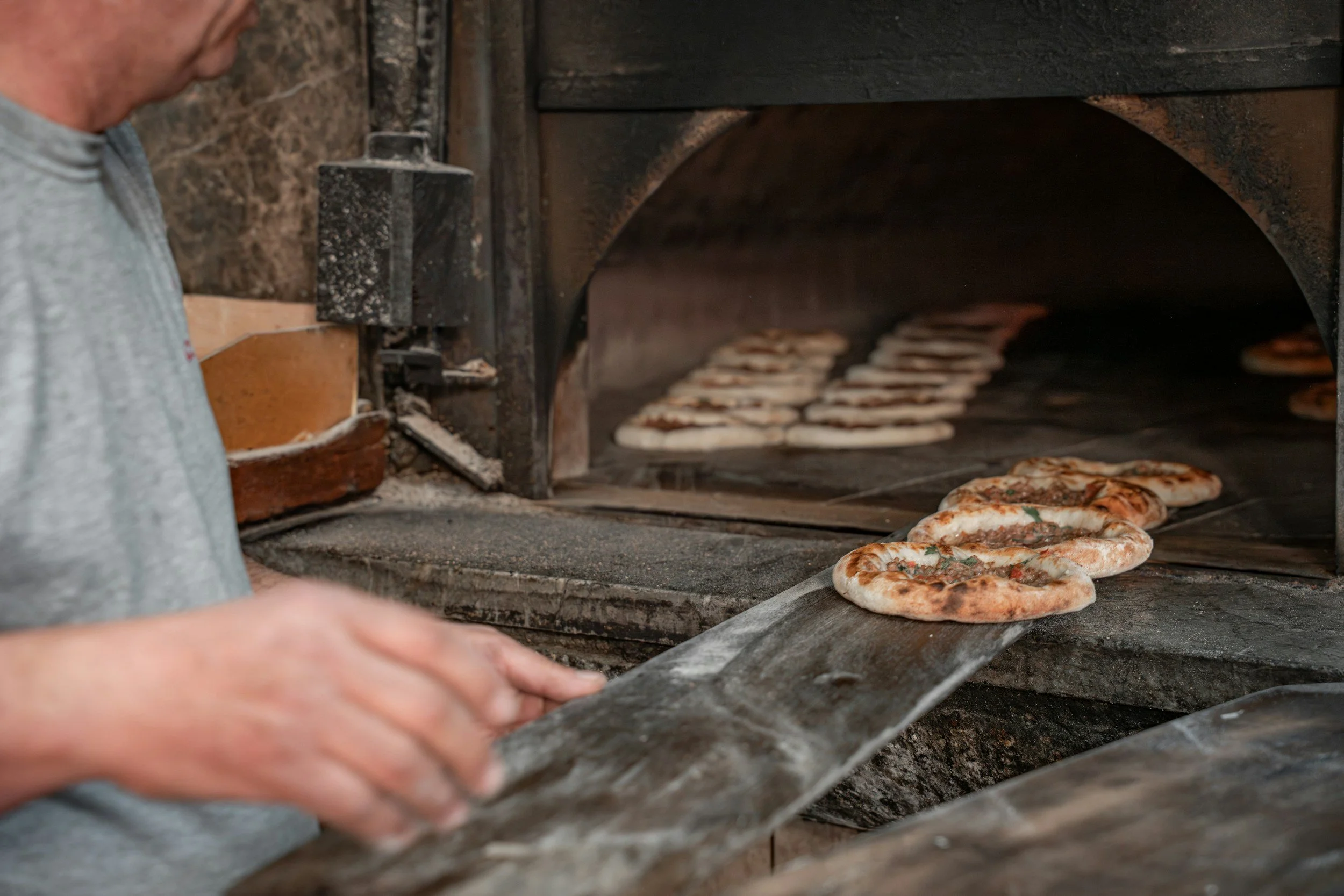 A person is baking pizzas in a brick oven, with several pizzas visible inside the oven and one being placed in the oven on a baking peel.