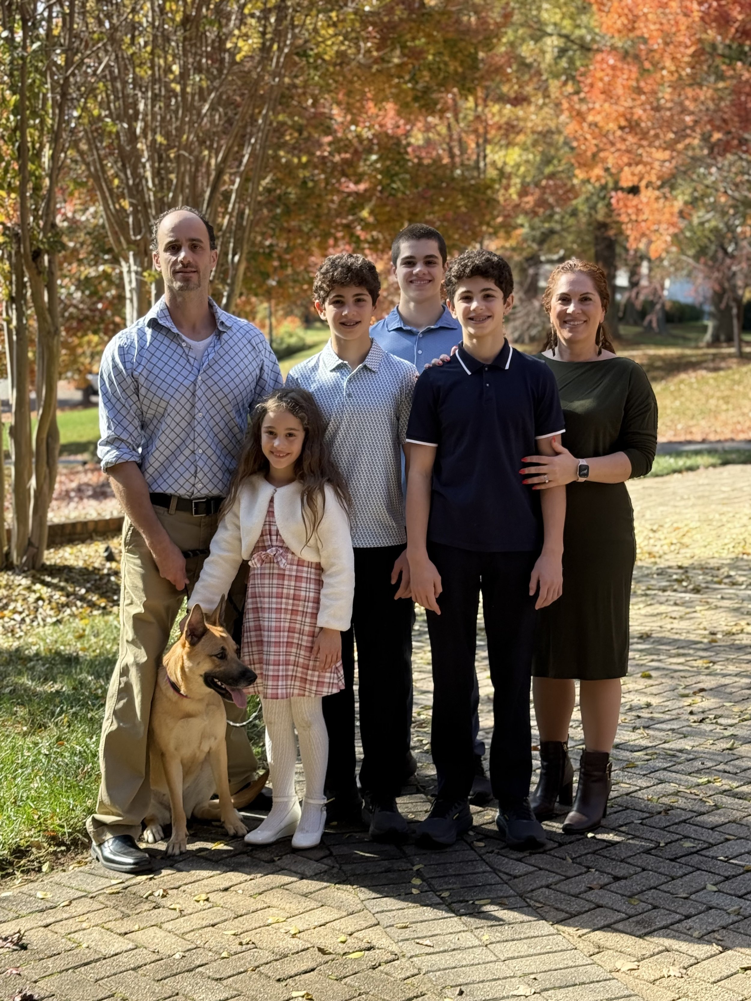 A family of seven with five children and two adults, including a dog, standing outdoors on a cobblestone path surrounded by trees with autumn leaves.