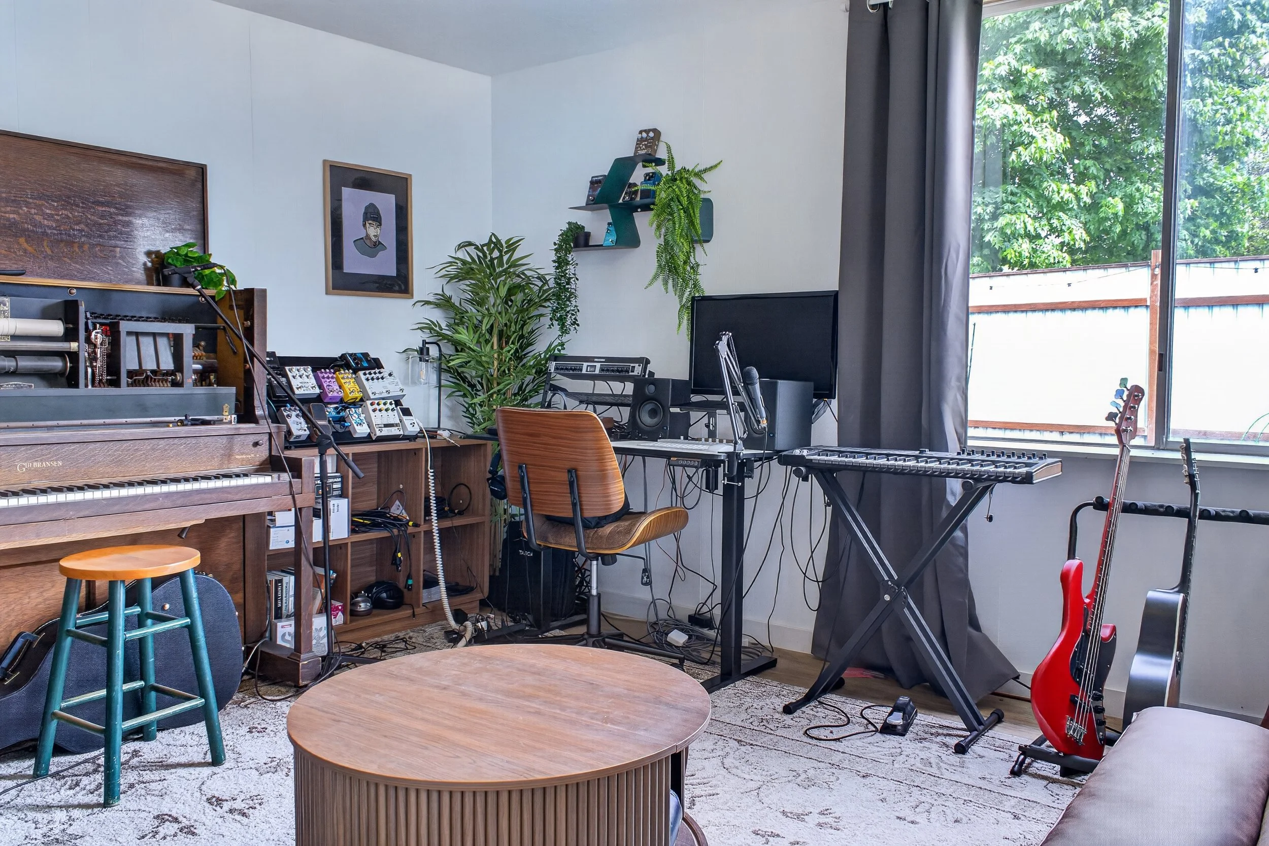 A music studio room with a upright piano, a variety of guitar pedals on a shelf, a desk with music equipment including a monitor and a keyboard, a red guitar leaning on the wall, a guitar on a stand next to the window, and various plants and decor.