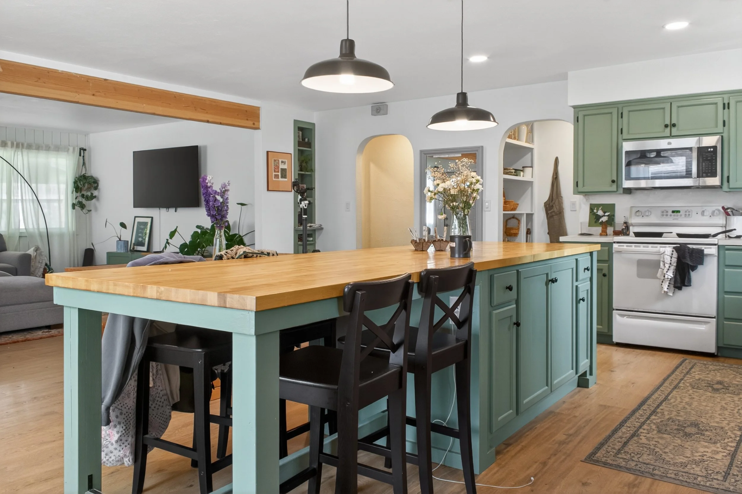 A kitchen with mint green cabinets, a wooden countertop island, black chairs, and stainless steel appliances. There are flowers on the island and a living room with a TV, curtains, and a couch in the background.