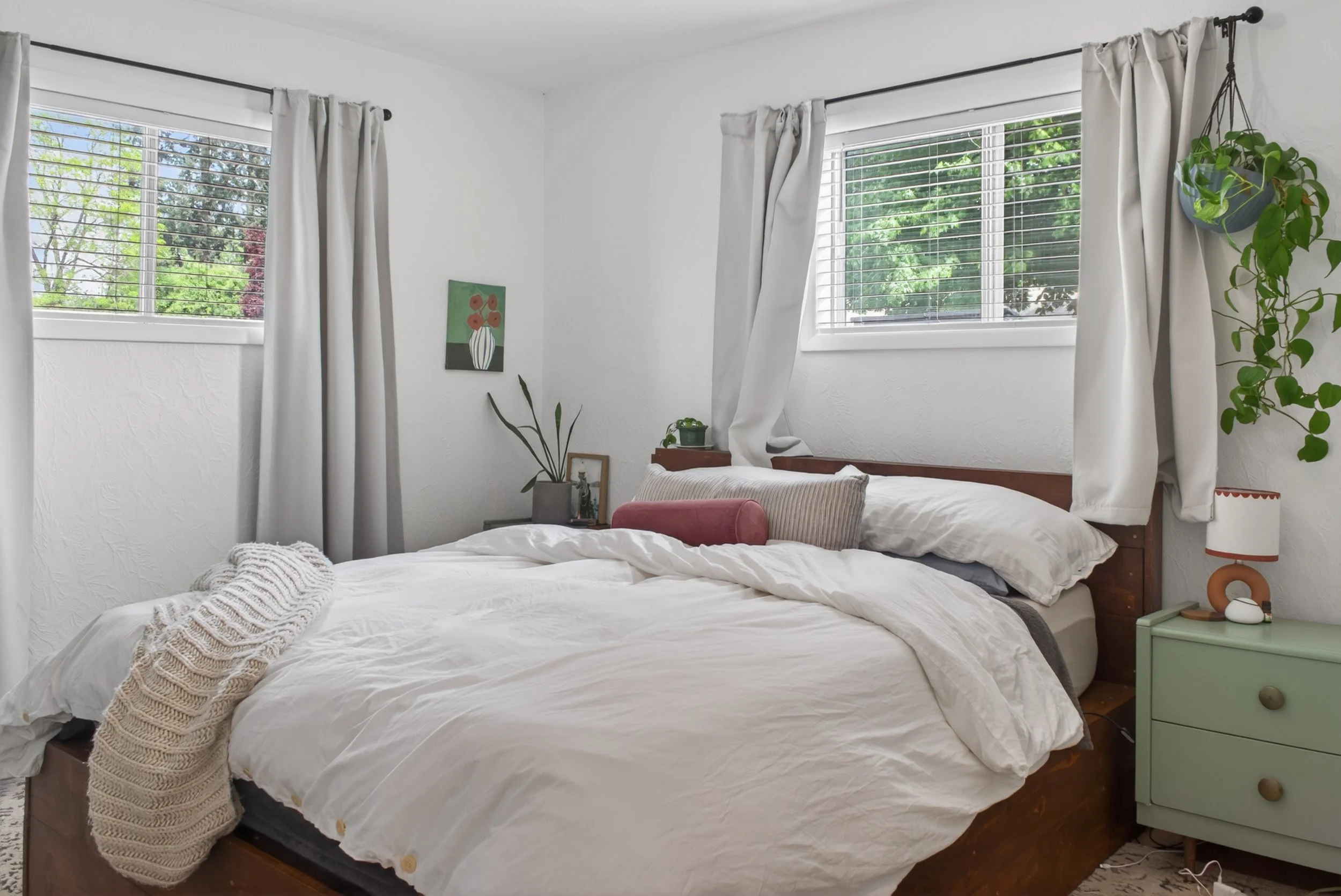 A bedroom with a wooden bed frame, white bedding, and a beige knitted blanket. There are two windows with white blinds and curtains, and a small green nightstand with a lamp and decor. Potted plants and a painting are on the wall.