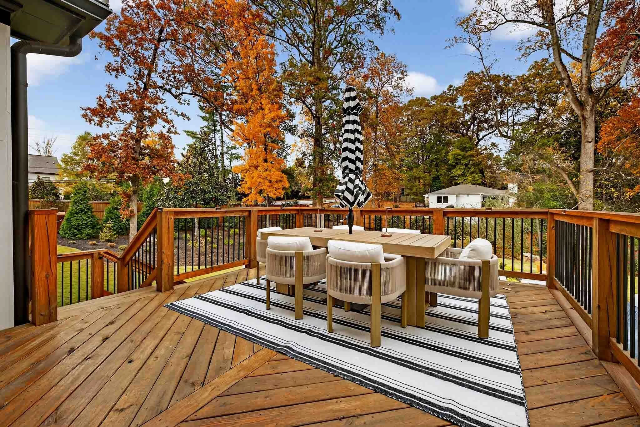 A backyard deck with outdoor furniture and a black-and-white striped patio umbrella, surrounded by trees with orange, yellow, and green leaves, indicating fall, with a blue sky in the background.