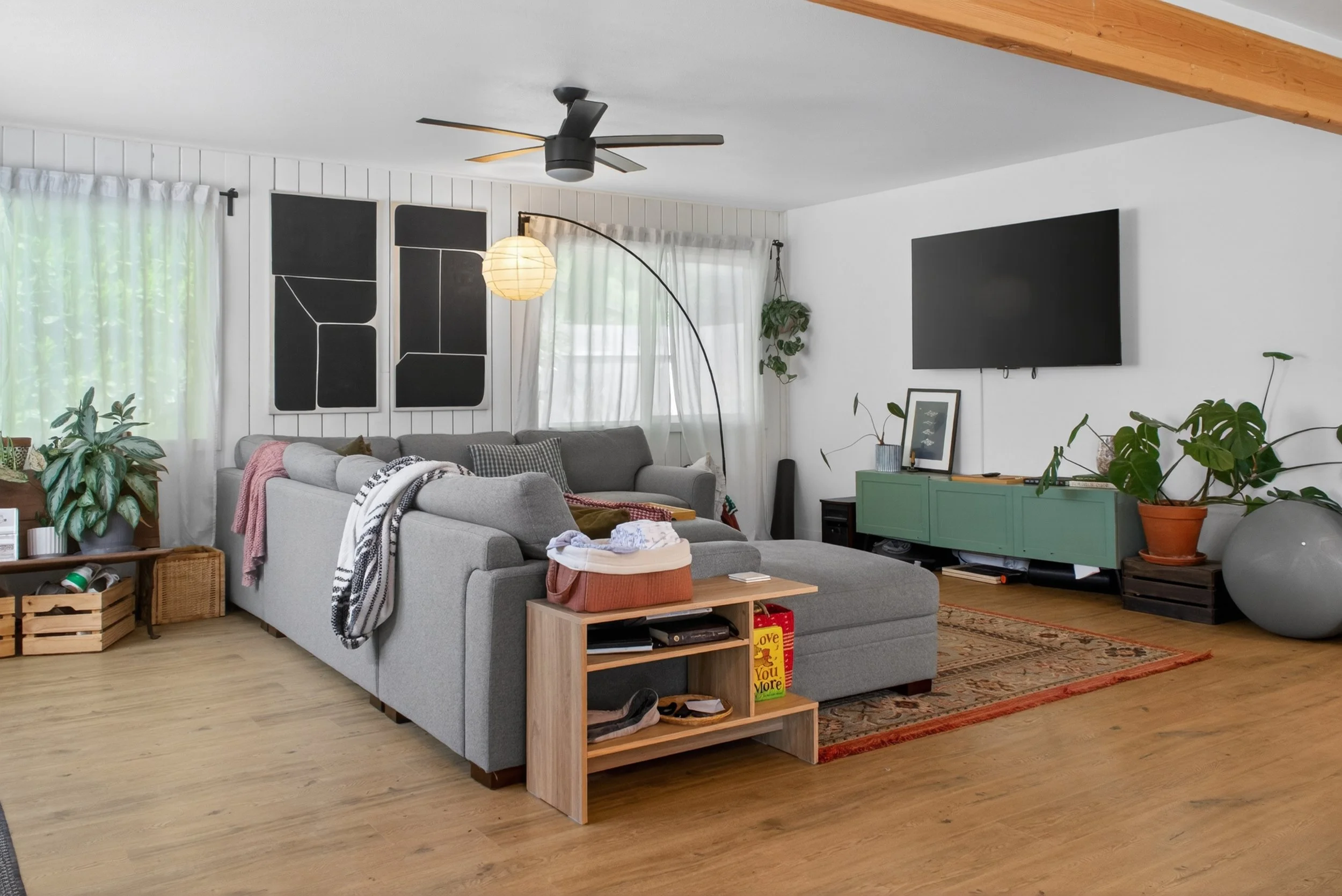 A living room with white paneled walls, a gray sofa with blankets and pillows, a wooden coffee table, a green cabinet, and various potted plants. There are windows with white curtains, wall art, a television, and a patterned rug on a hardwood floor. A black ceiling fan is on the ceiling, and a modern arc floor lamp with a paper lampshade is next to the sofa.