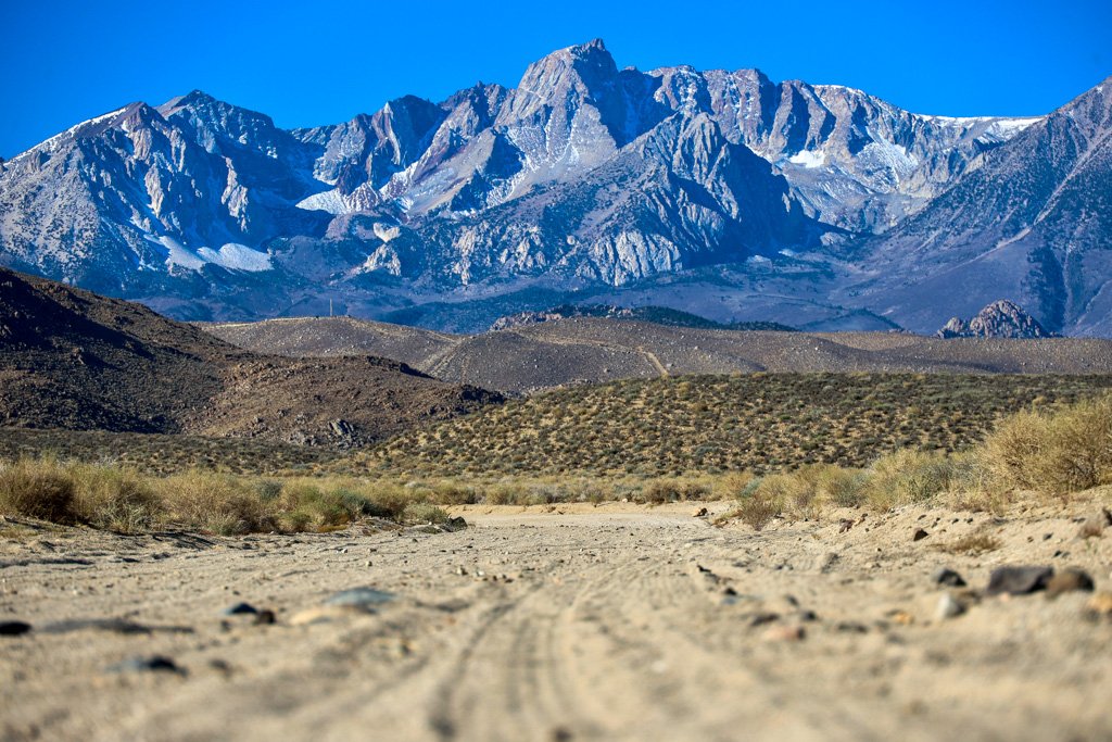 A desert trail leading to rugged mountains with snow-capped peaks under a clear blue sky.