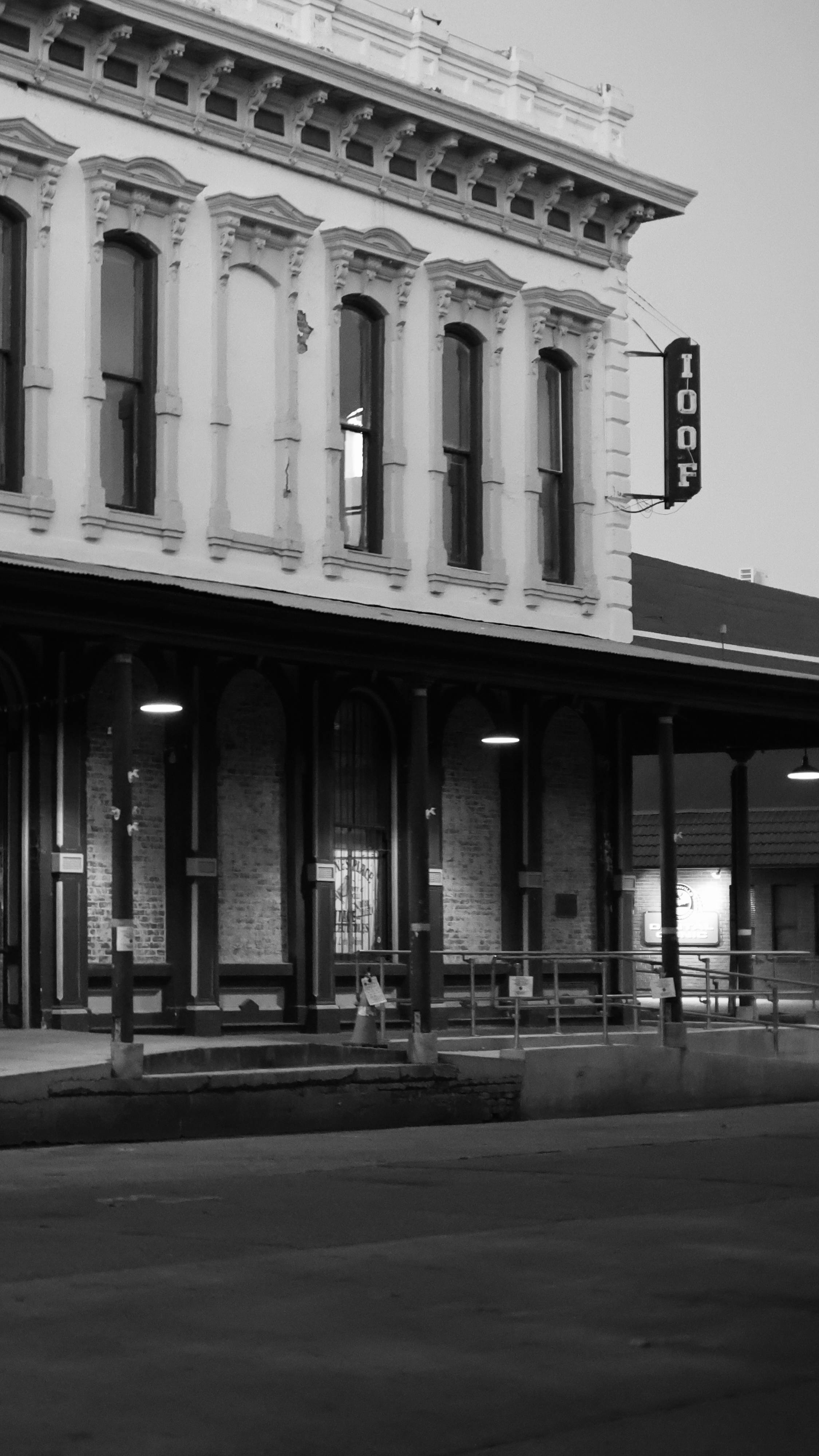 Black and white photo of a two-story building with ornate architectural details, large windows, and a vertical sign that reads 'TOOF' hanging from the corner. There is a covered sidewalk area underneath with hanging lights and concrete barriers along the street.