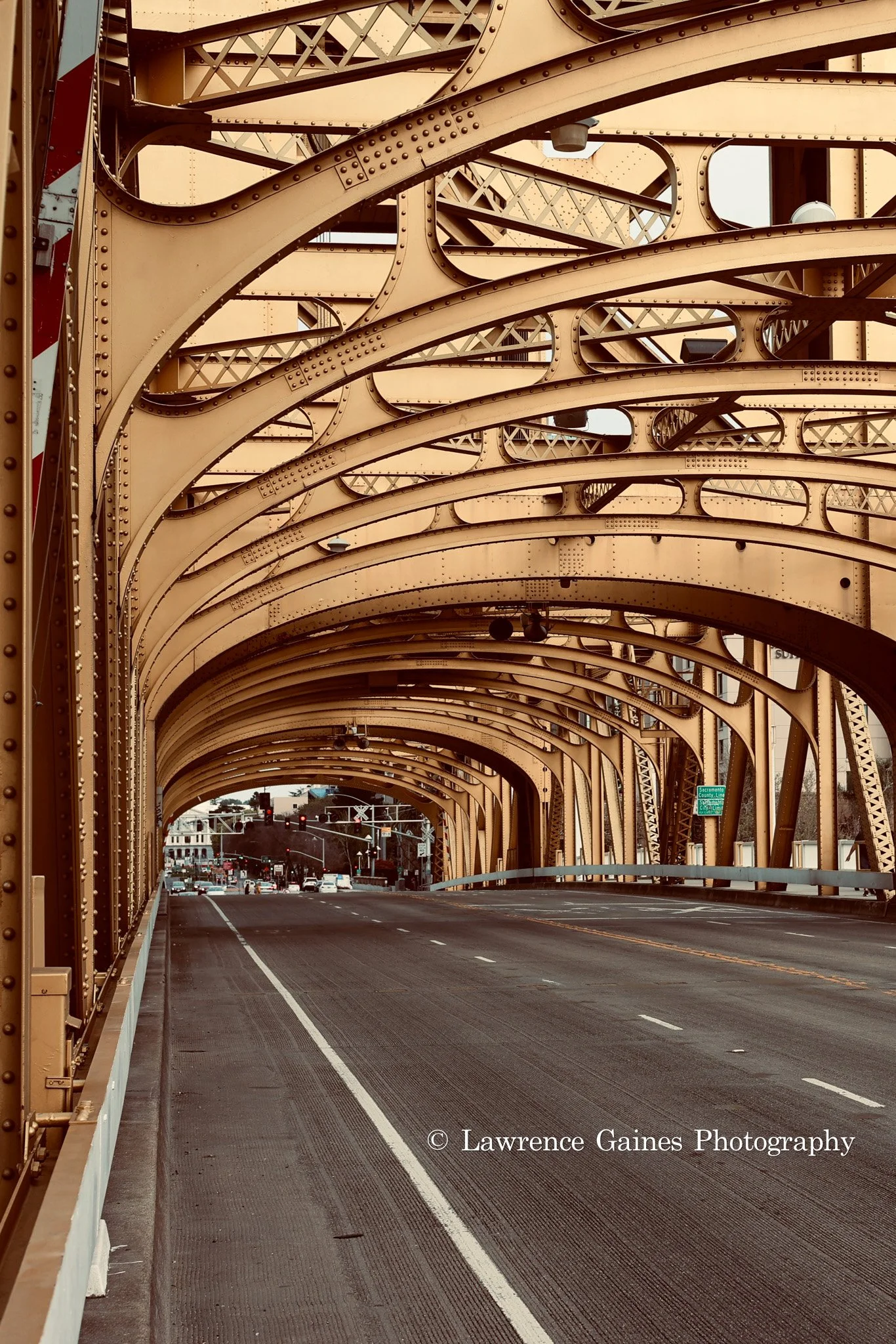 Multiple yellow metal arches form the structure of a bridge, with the road underneath mostly clear of traffic, leading to traffic lights and city buildings in the background.