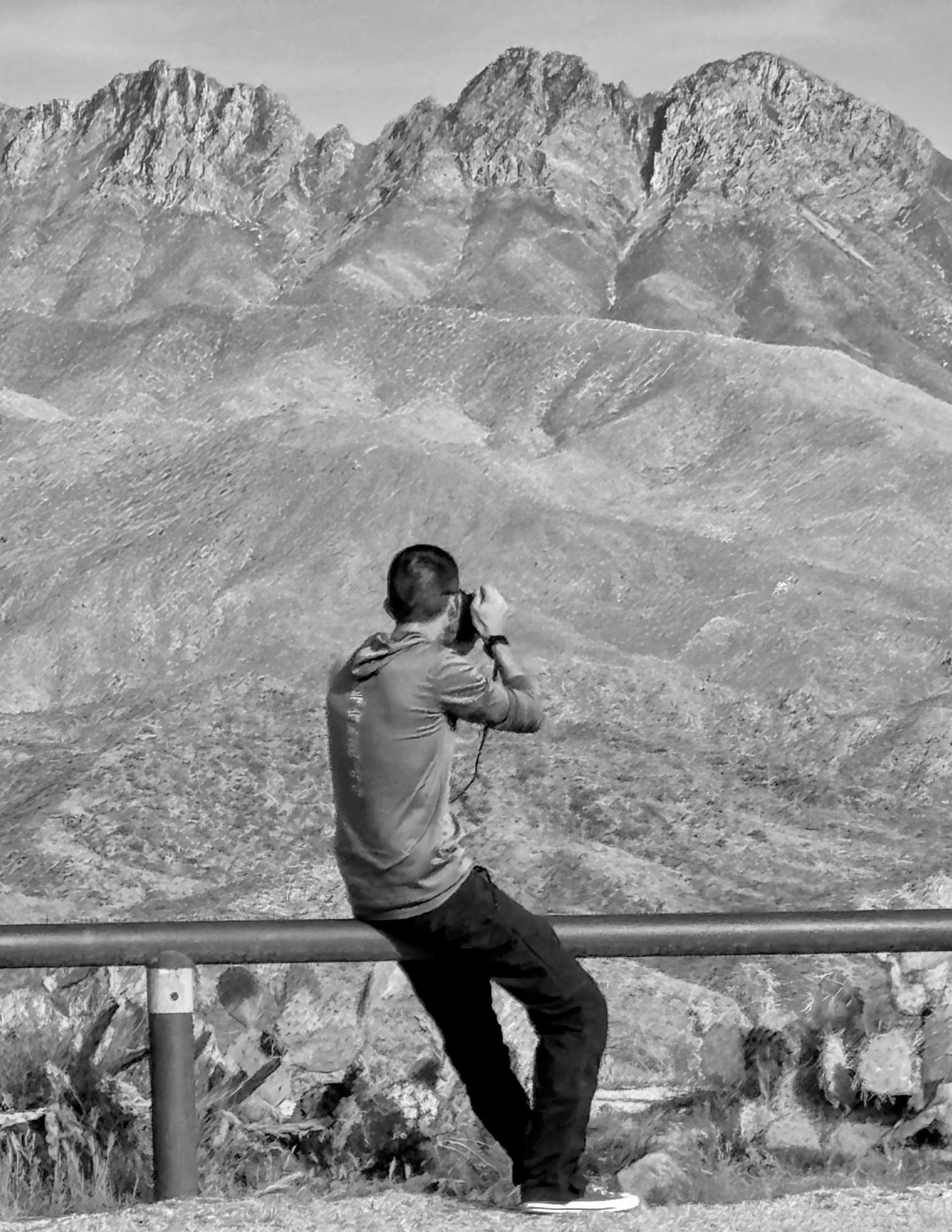 Black and white photo of a boy in a jacket and jeans, standing outdoors, with mountains in the background. The boy appears to be looking away and holding something in his hand.