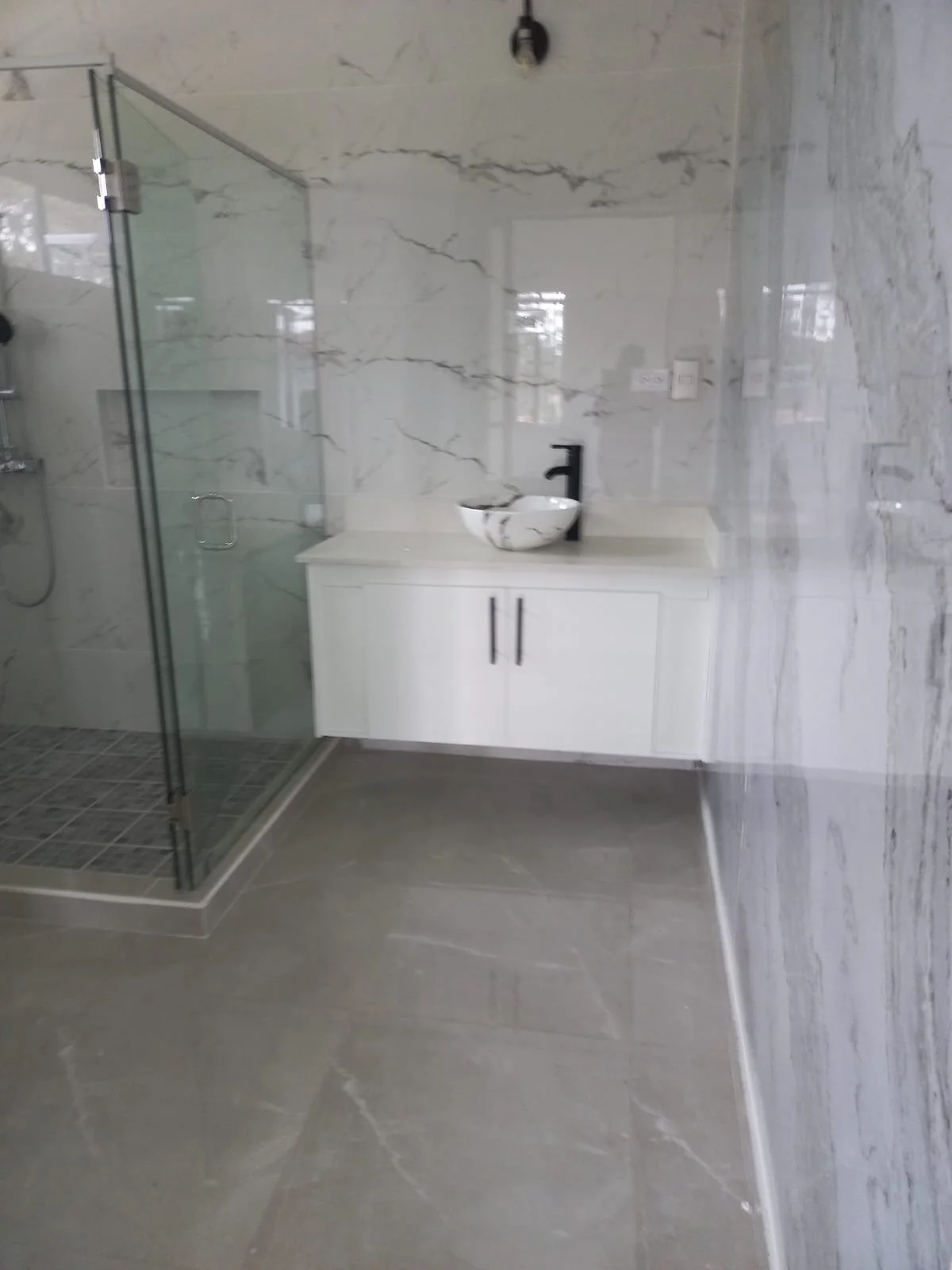 Modern bathroom with marble walls and floor, a walk-in shower with glass door, and a white vanity with a vessel sink and black faucet.