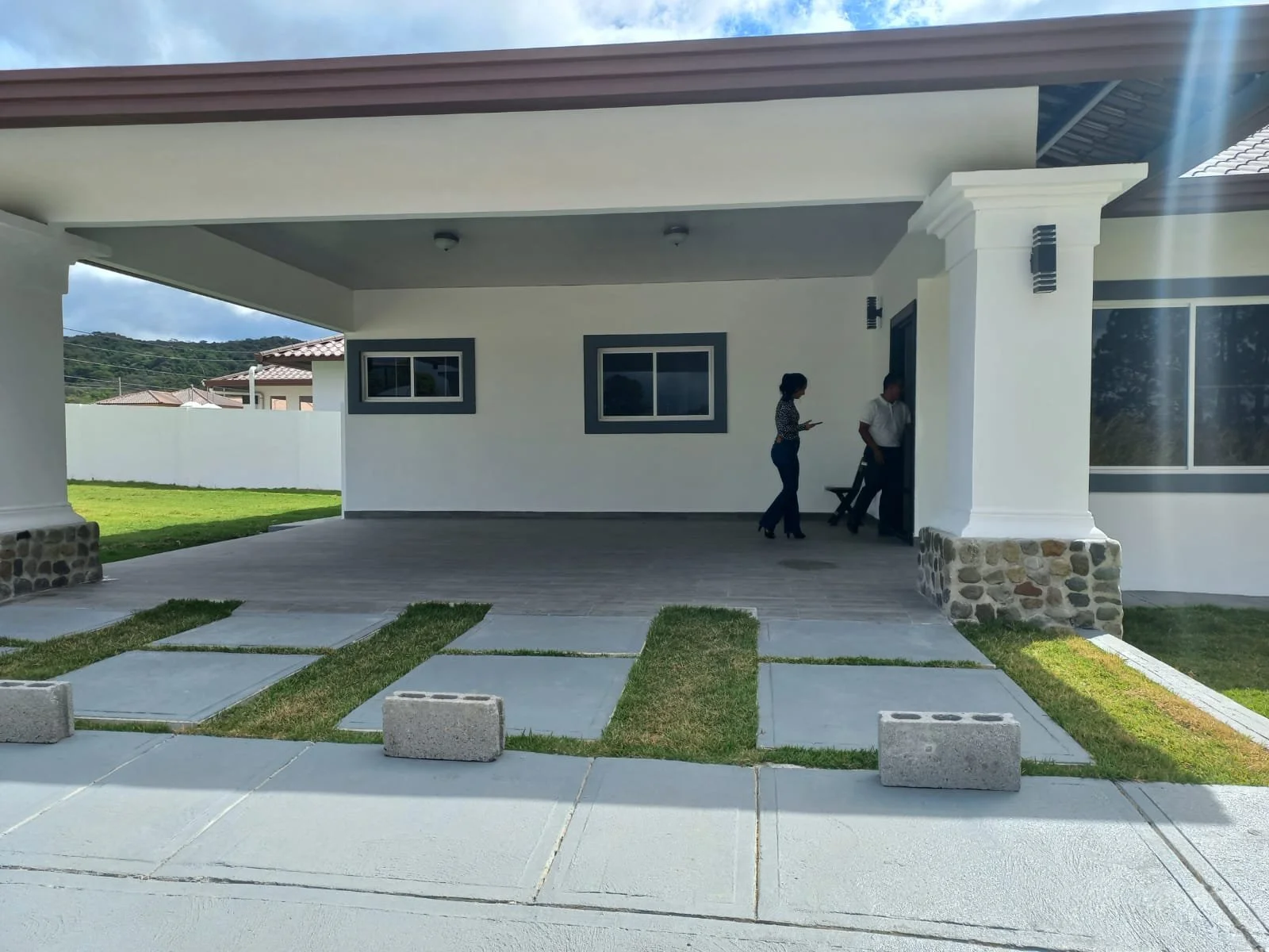 People standing at the entrance of a modern house with a covered porch, stone accents, and a front yard with concrete stepping stones and grass.