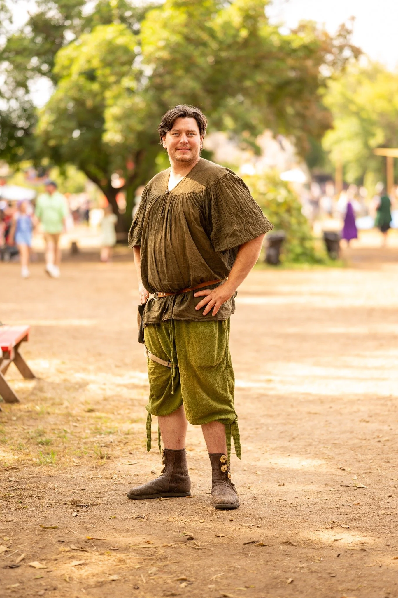 A man dressed in a medieval or fantasy-style costume, standing outdoors on a dirt path in a park with trees and blurred people in the background.