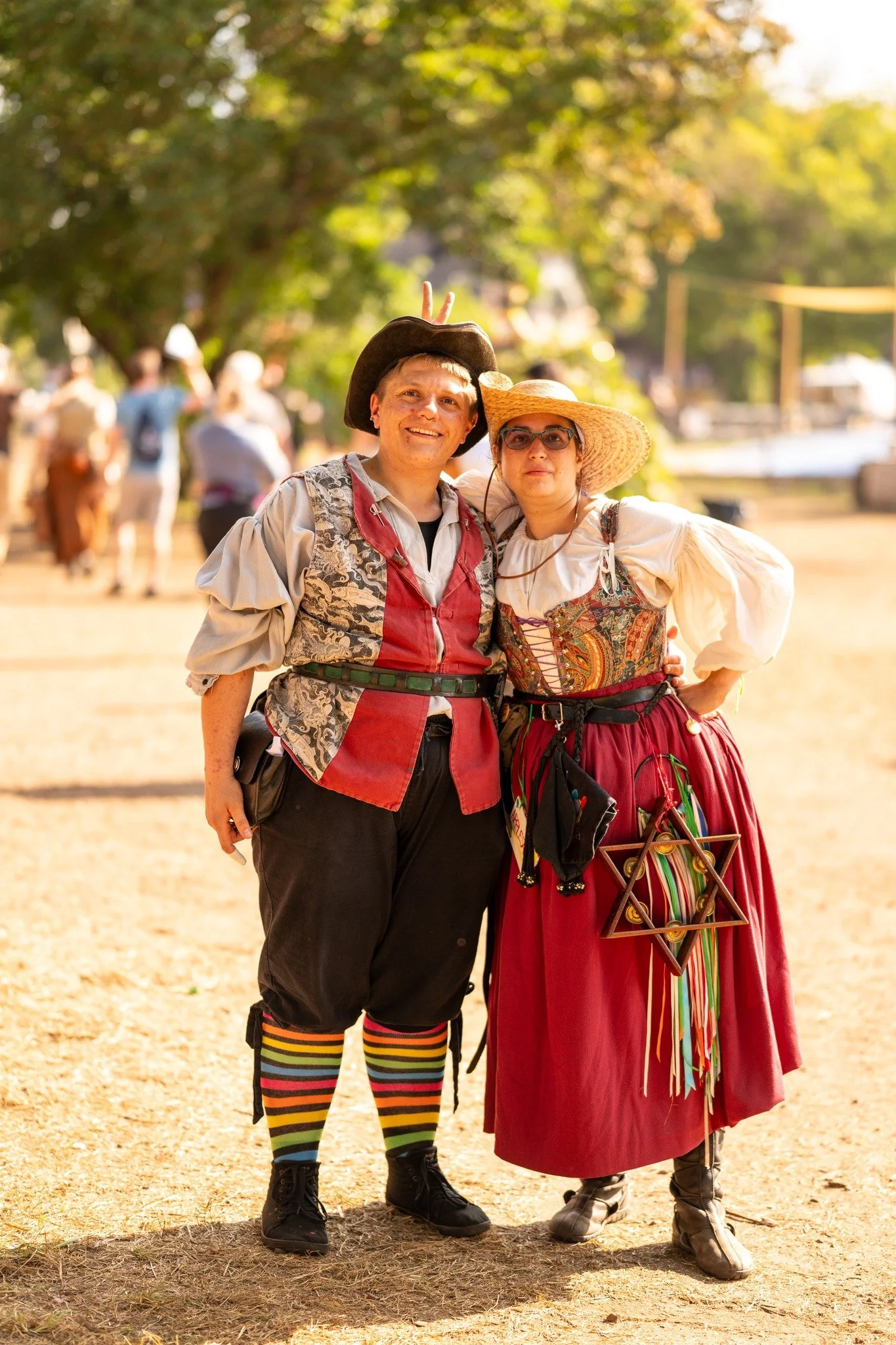 Two people dressed in traditional German costumes, standing outdoors on a sunny day, with trees and other people in the background. One wears a black hat and a waistcoat, and the other wears a wide straw hat and a long skirt.