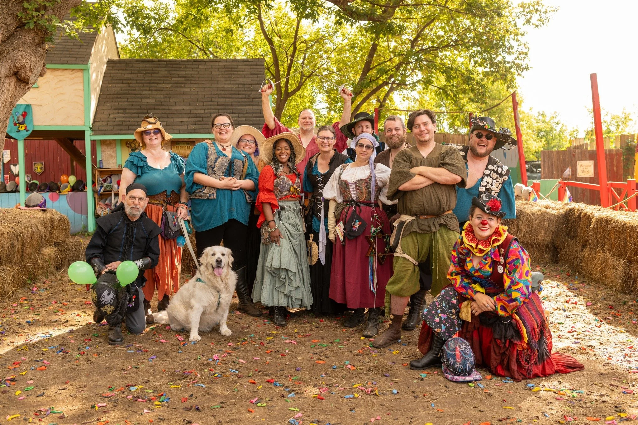 Group of people dressed in Renaissance costumes at an outdoor festival, with a dog and balloons, surrounded by hay bales and trees.