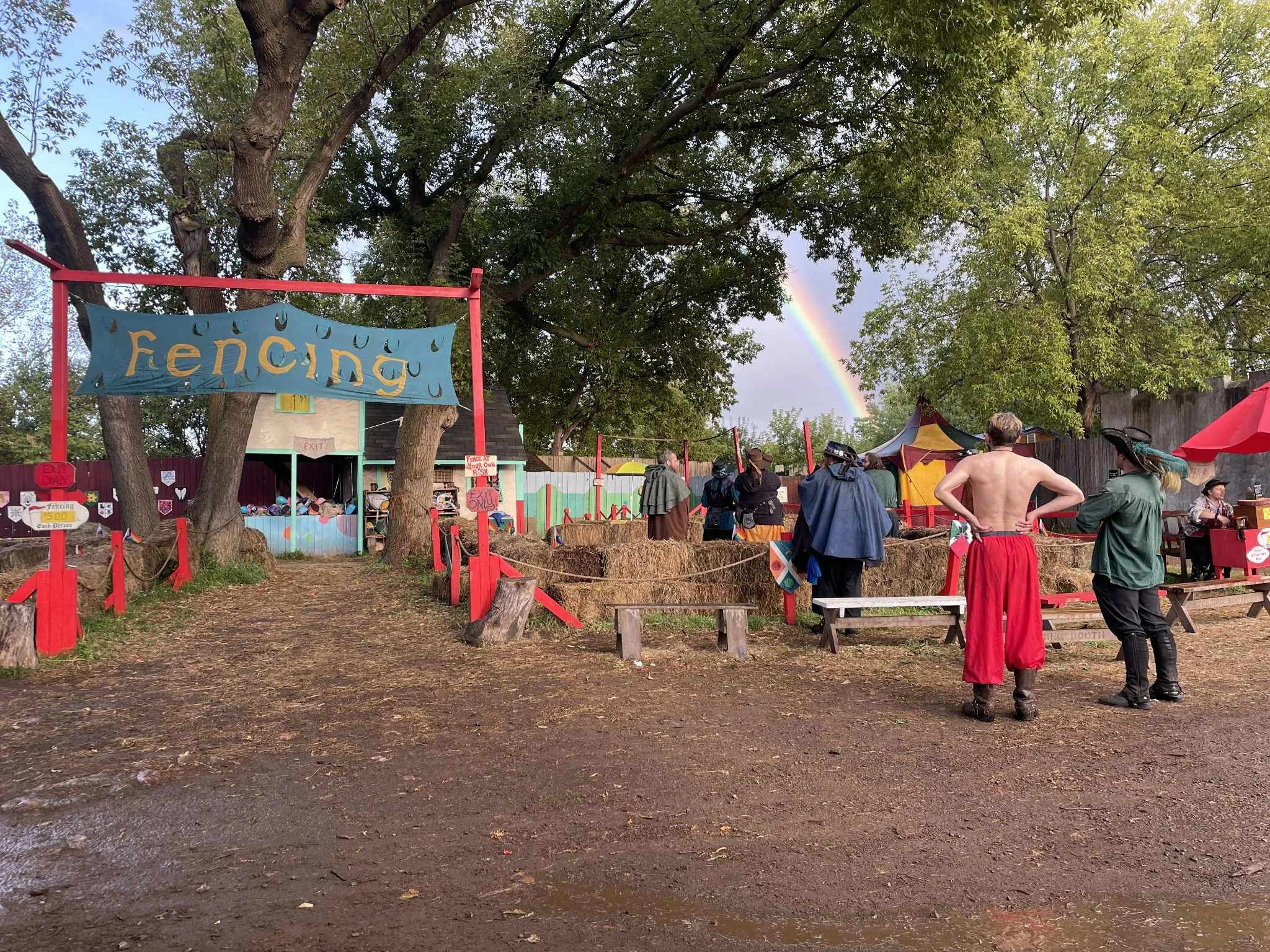 An outdoor scene at a festival with people wearing medieval or fantasy costumes, large trees, a rainbow in the sky, and colorful tents and signs.