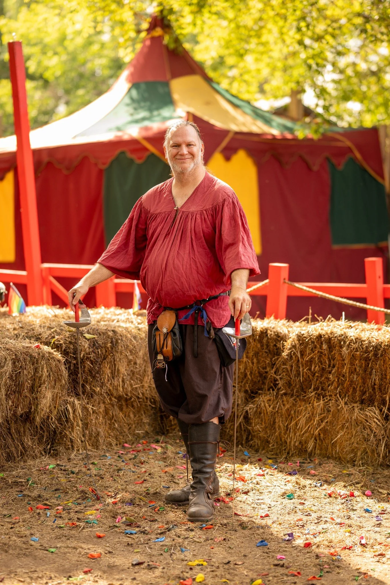 A man dressed as a medieval or fantasy knight, wearing a red tunic and chainmail, standing in a festive outdoor setting with a colorful circus tent in the background. The ground is decorated with multicolored confetti, and the scene is surrounded by hay bales.