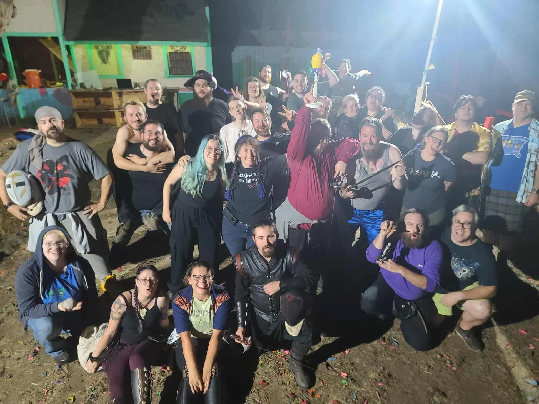 Group of people posing outdoors at night, some making peace signs, with colorful tents and decorations in the background.