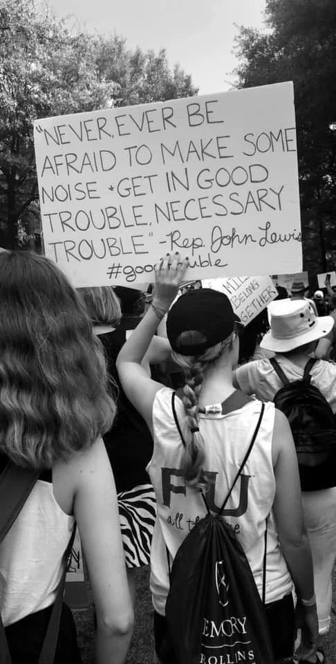 Group of people at a protest holding signs, one sign features a quote by Rep. John Lewis about bravery and making noise.