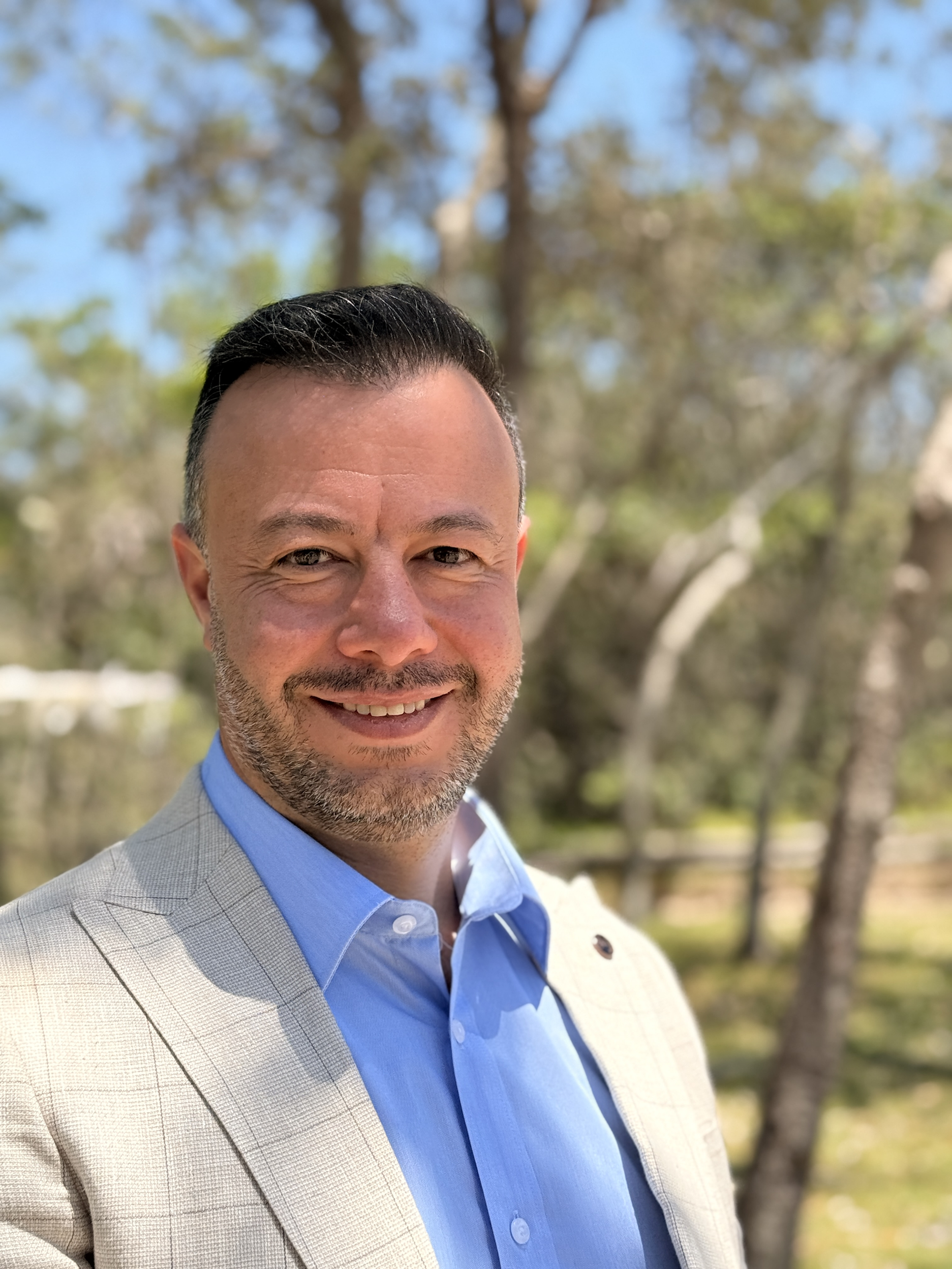 A smiling man in a light gray checkered suit with a blue shirt standing outdoors near trees.