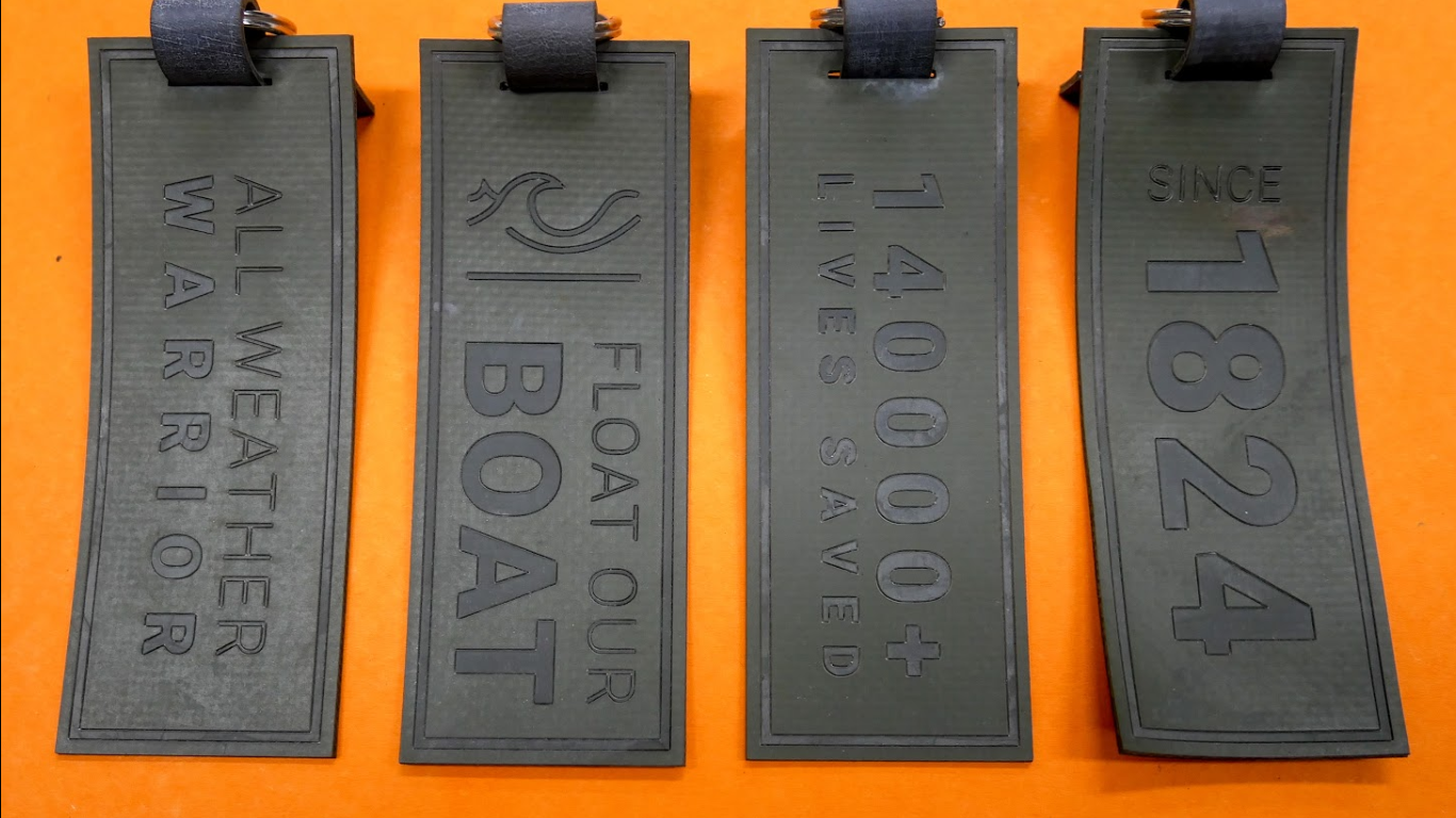 Four black rubber price tags with embossed text and numbers, attached to loops, displayed on an orange surface. The tags feature inscriptions such as "ALL WEATHER WARRANTY," "FLOAT OUR BOAT," "$4000+ LIVEST SAVED," and "SINCE 1824."